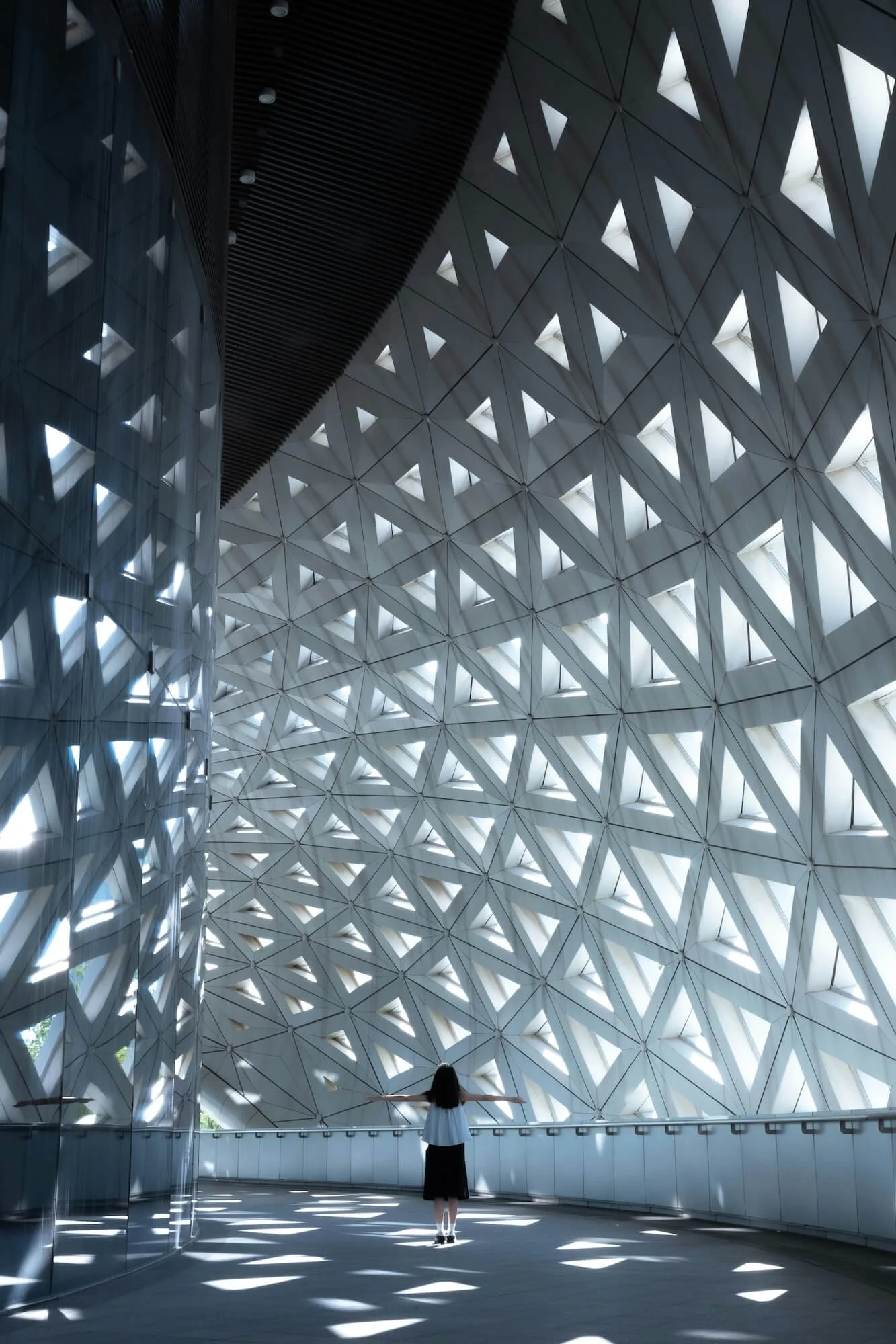 A person standing inside a modern architectural structure with a large patterned wall made of triangular cutouts allowing light to shine through.