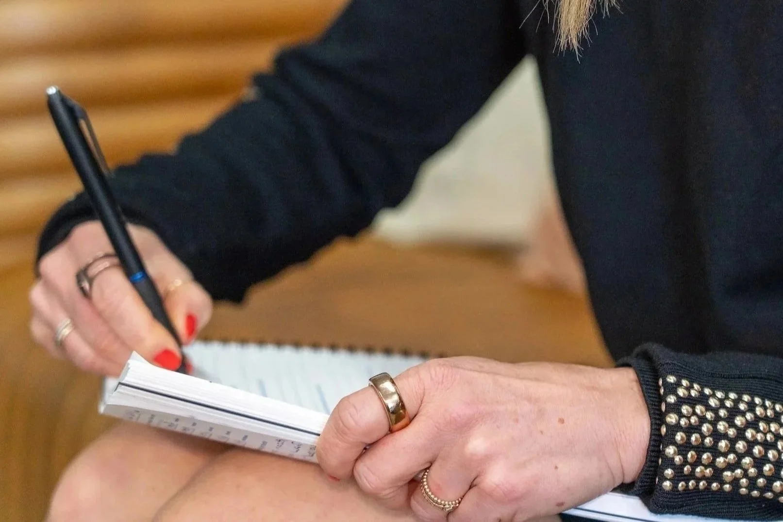 Person taking notes in a notebook with a pen, wearing rings and a black sweater with silver studs on the sleeves.