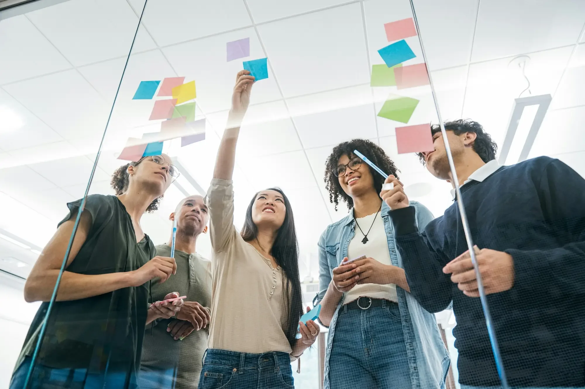 Five diverse people in a meeting room with glass walls, collaborating and placing colorful sticky notes on the glass surface for a planning or brainstorming session.