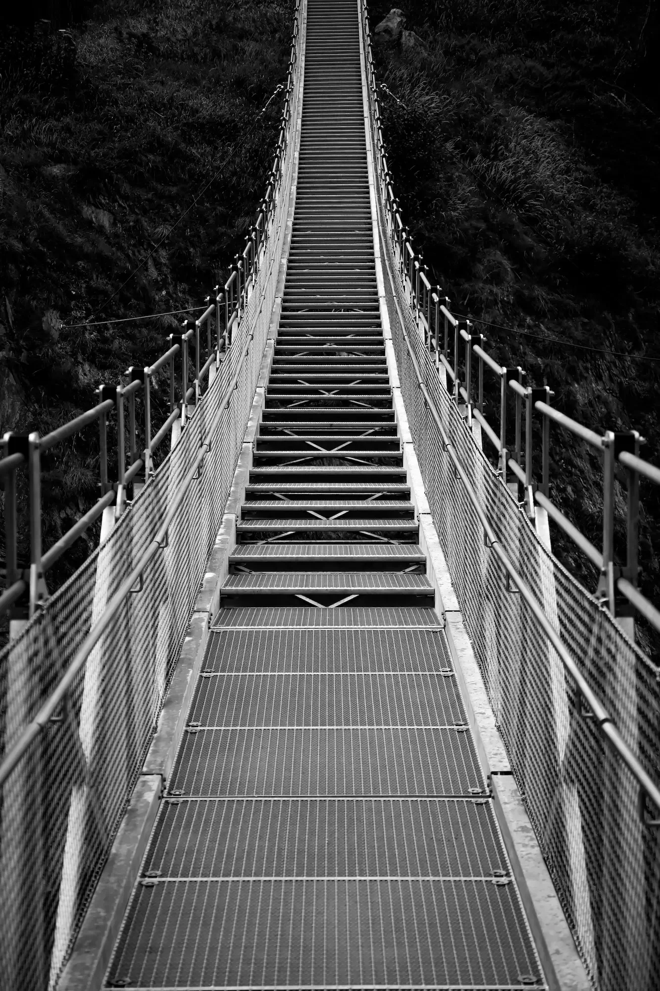 A long suspension bridge with metal grating flooring stretches upward, flanked by metal railings on both sides, over a dark, rocky landscape.