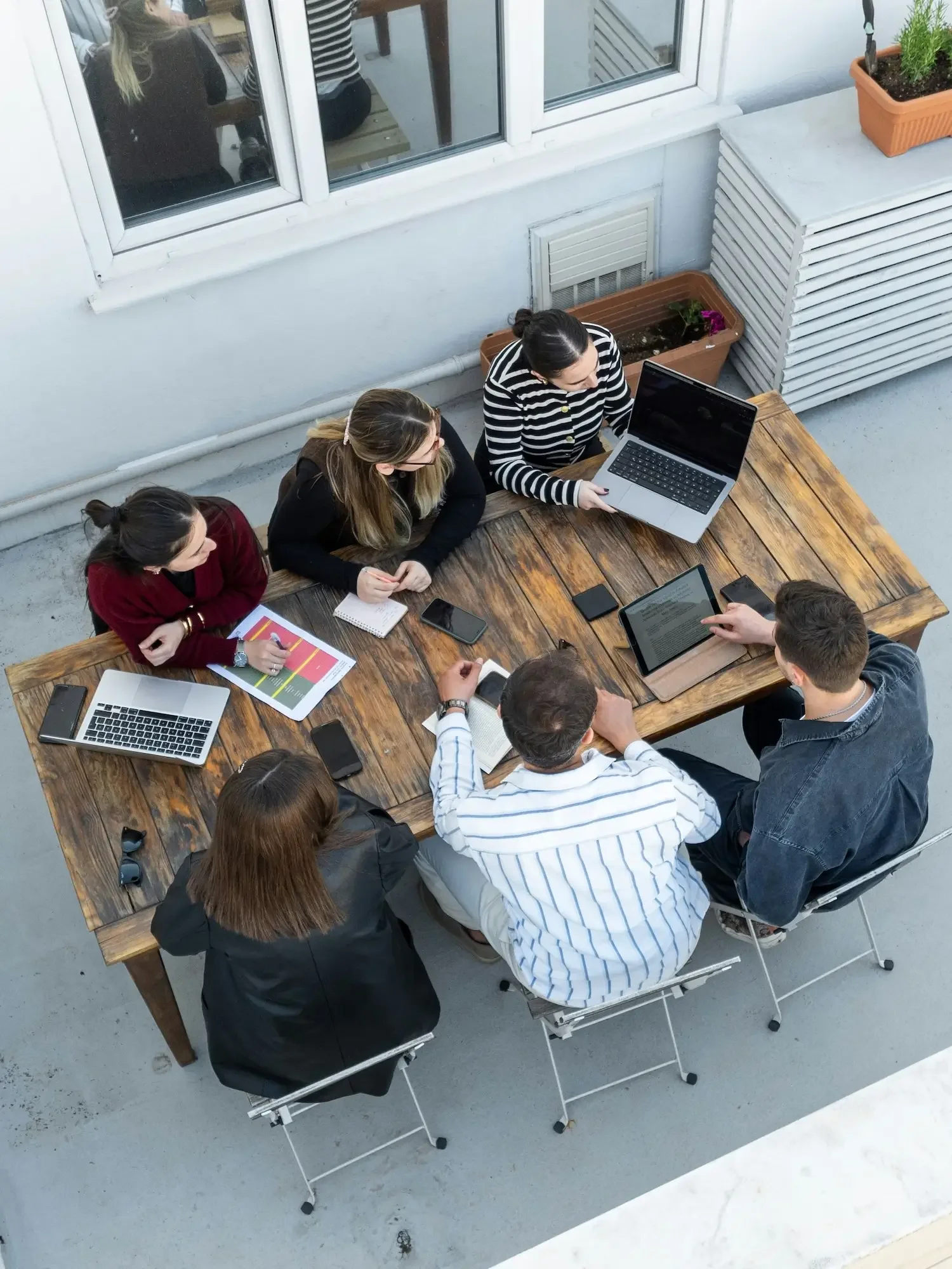 Six people sitting around a wooden table on a balcony, working with laptops, tablets, and smartphones, with some papers and a notebook.