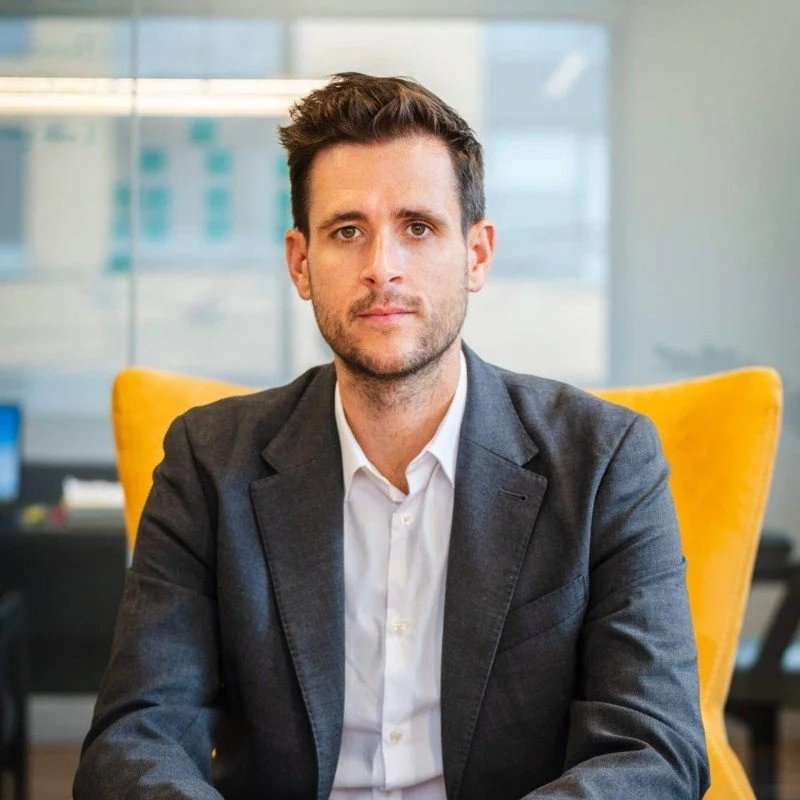 Portrait of a young man with brown hair, beard, wearing a dark blazer and white shirt, sitting in a yellow chair in an office setting.