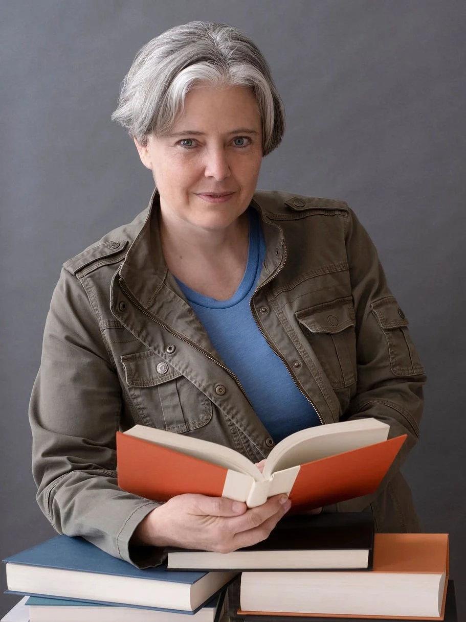 The book midwife Fen Druadìn leans on a stack of book while holding an open book and looking at the viewer.
