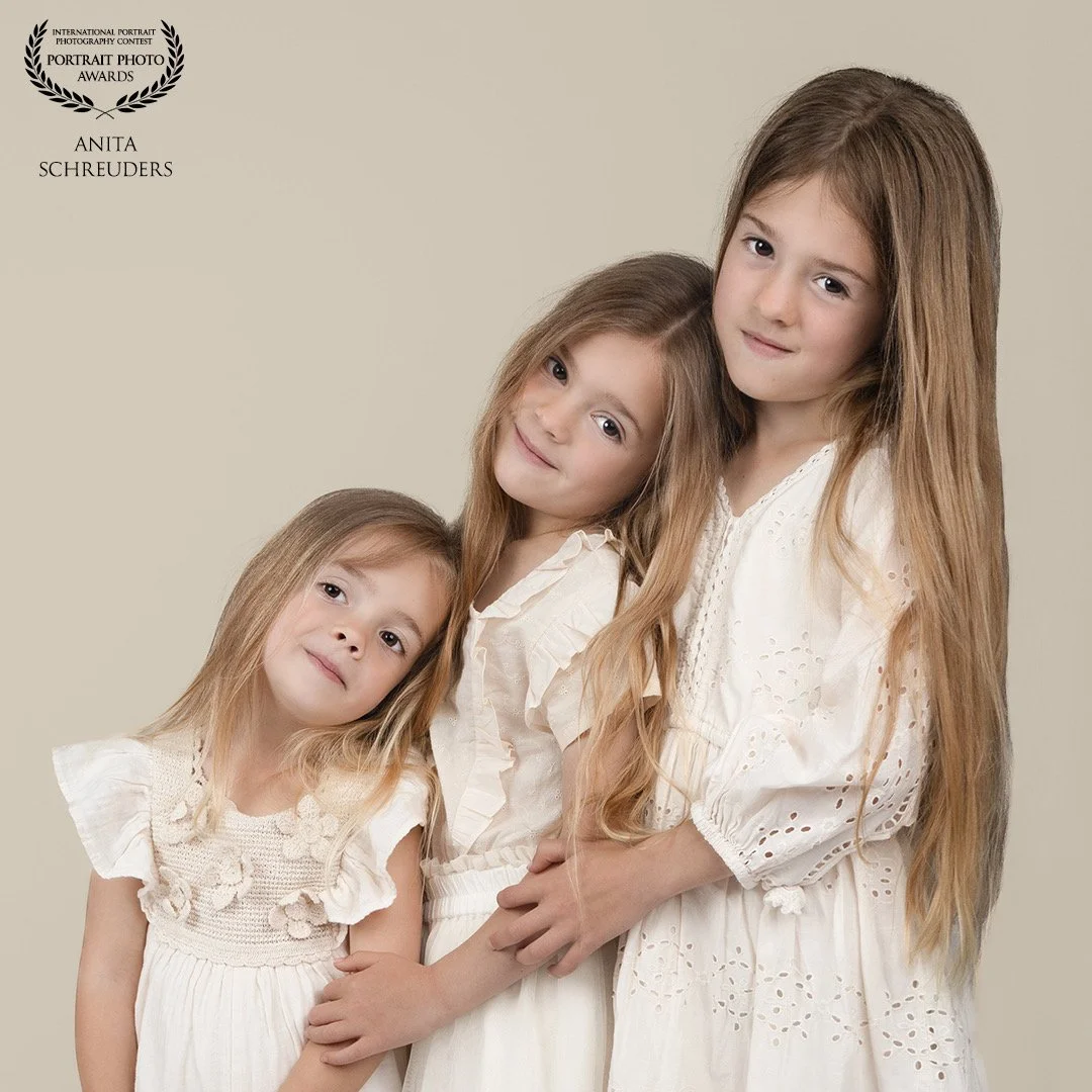 Drie jonge meisjes met lang, lichtbruin haar in witte kleding staan dicht bij elkaar, poseren voor een neutrale achtergrond.