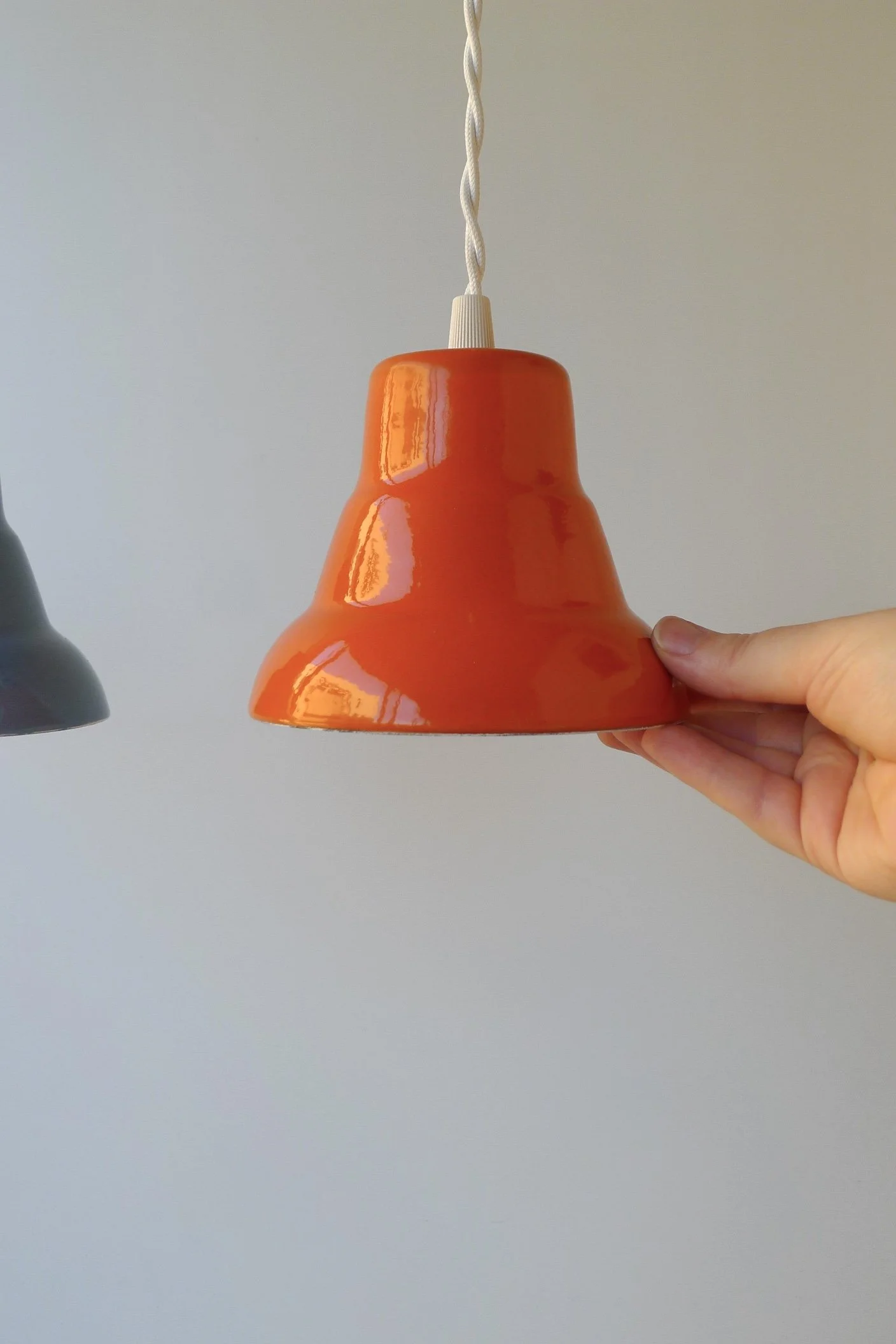 Bright orange pendant light with a glossy finish, held by a hand. The light fixture has a bell-shaped shade and a white cord. A second, darker pendant is partially visible on the left.