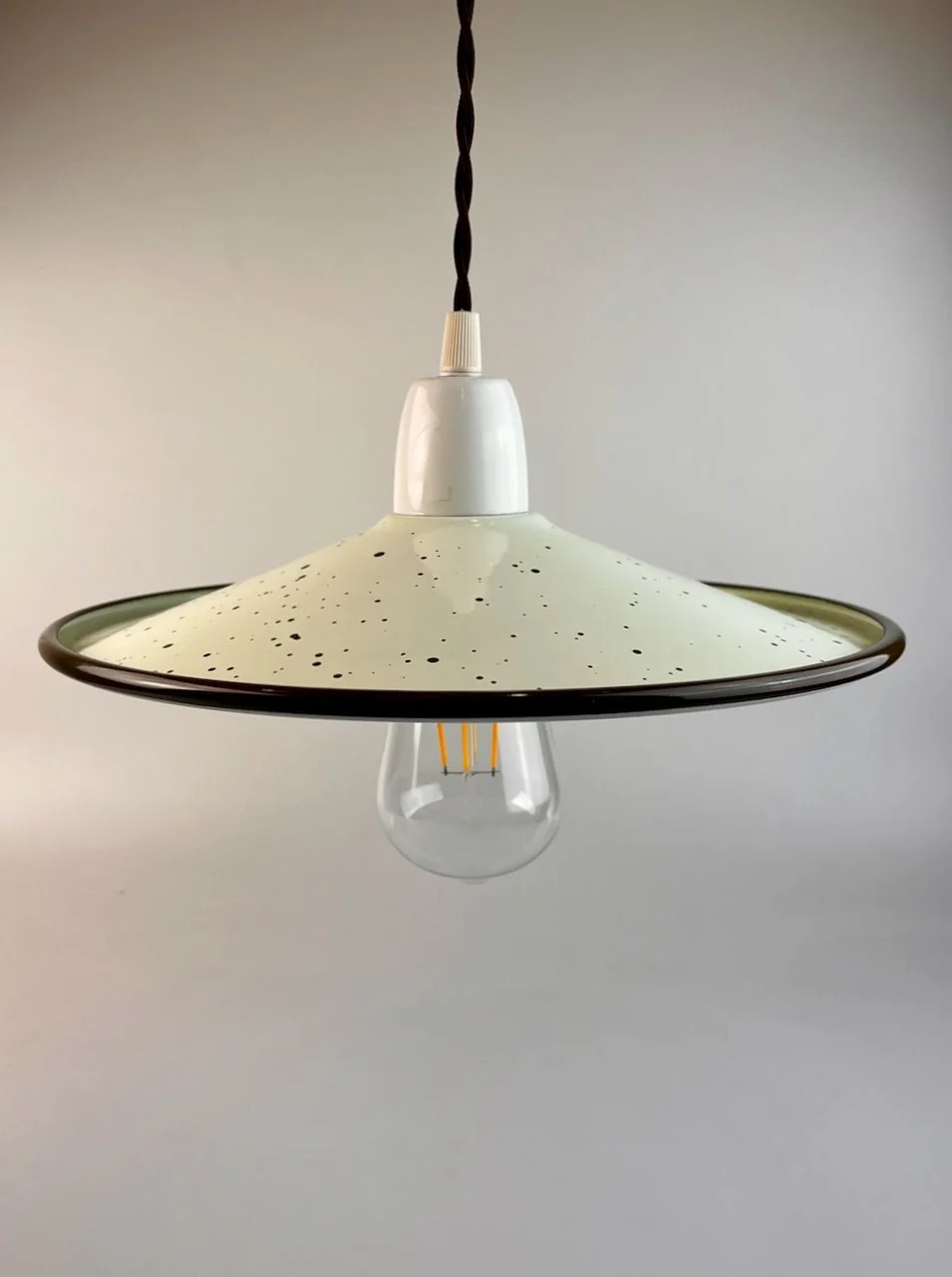 Pendant light fixture with a cream, speckled, cone-shaped shade and exposed light bulb, hanging from a twisted dark cord.