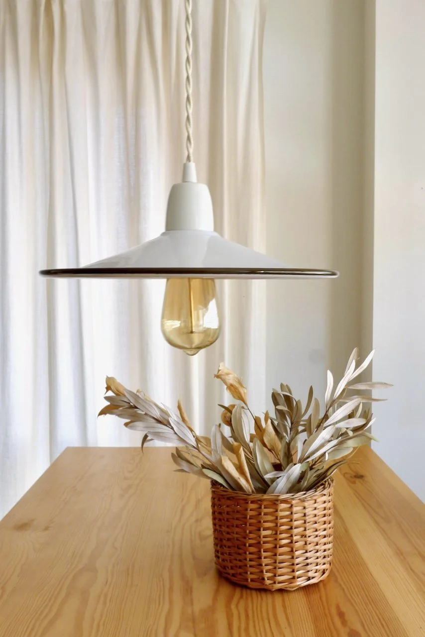 A hanging white pendant light with a visible light bulb, placed above a wooden table with a wicker basket filled with dried beige and light brown leaves, in front of cream-colored curtains.