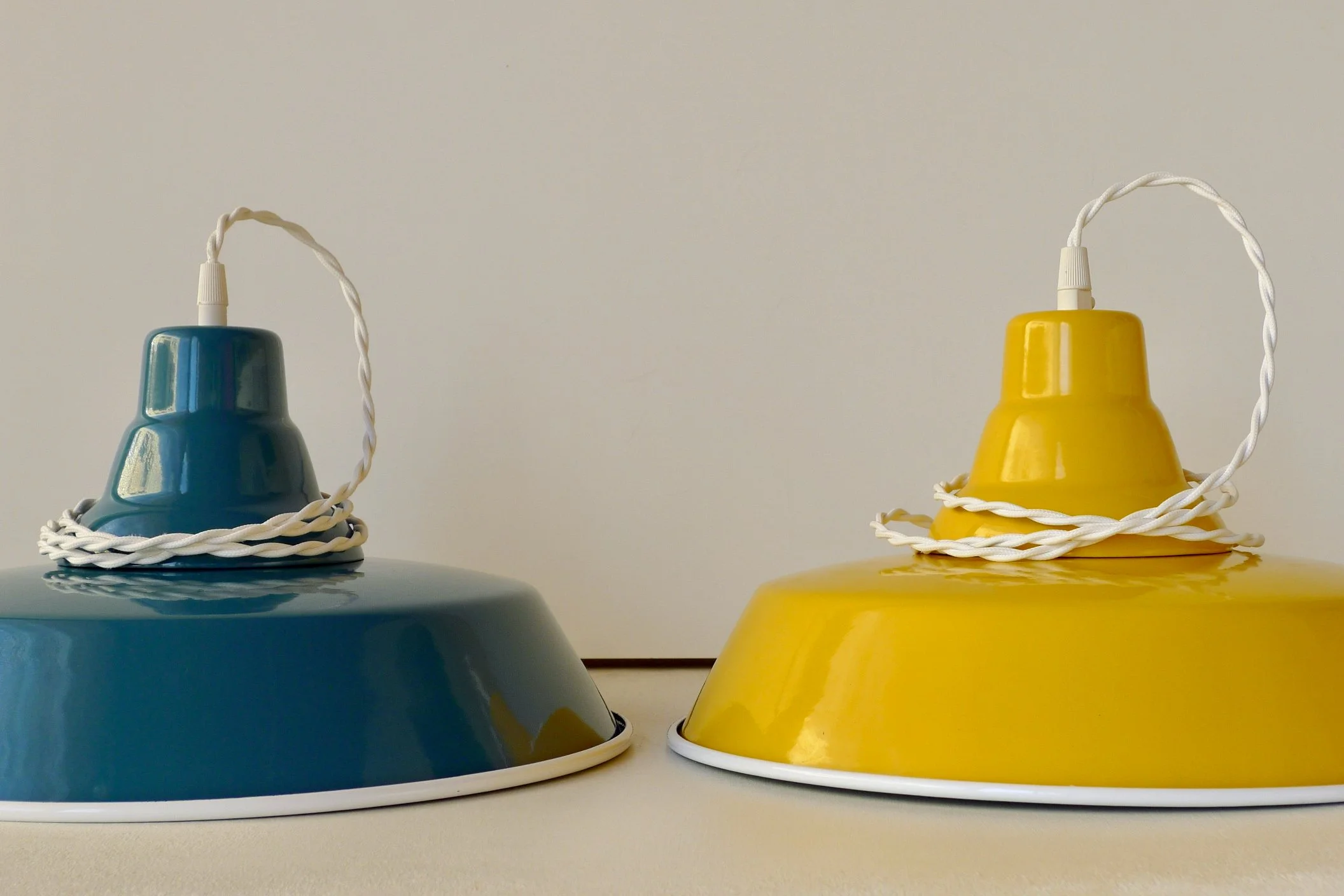 Two enamel table lamps, one green and one yellow, with twisted white cords, placed on a white surface against a plain white background.
