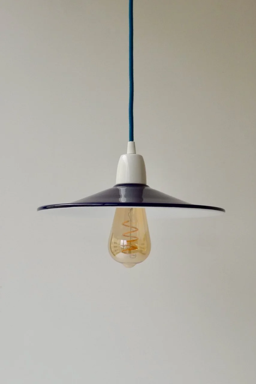 A pendant light fixture with a navy-blue shade, white holder, and an exposed filament bulb hanging from a petrol-blue cord against a plain wall.