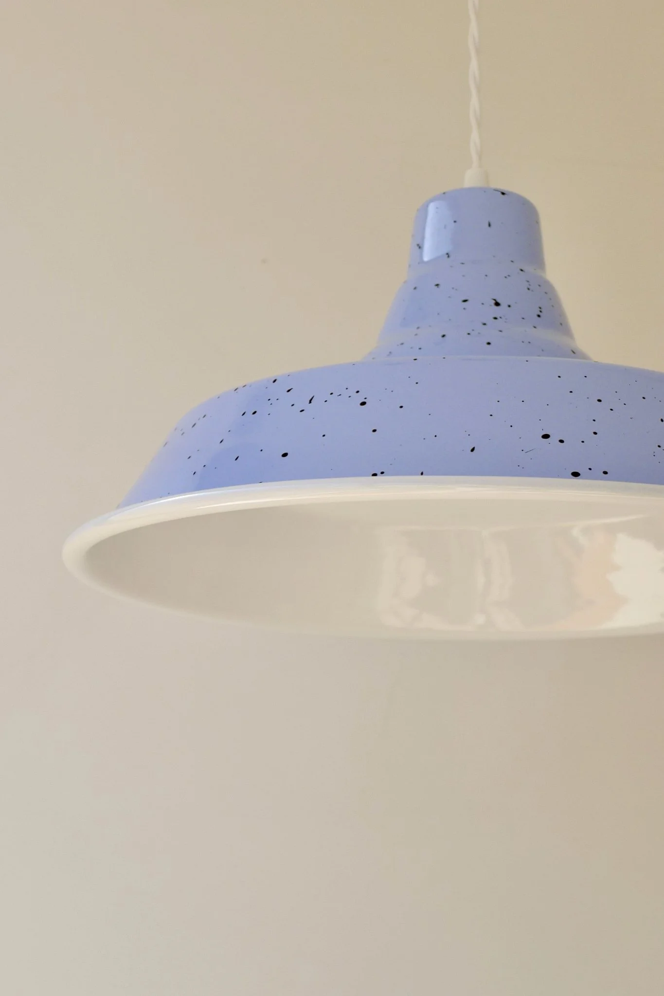 Close-up view of a lavender and white ceiling pendant light with dark-brown speckles, hanging from a white twisted cord against a plain beige ceiling.