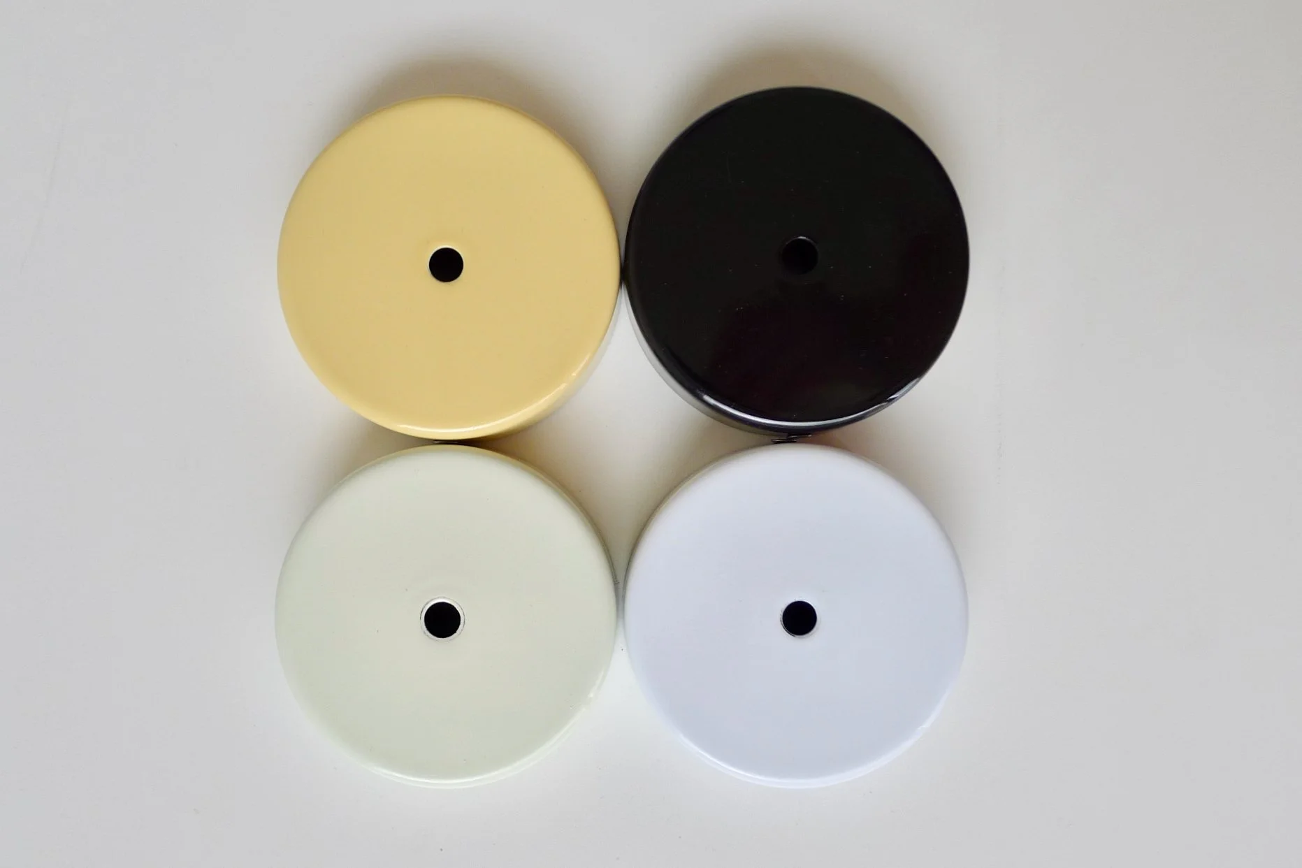 Four circular flat ceiling roses with holes in the center arranged in a 2x2 grid on a white surface. The enamel roses are black, white, cream, and light-yellow.