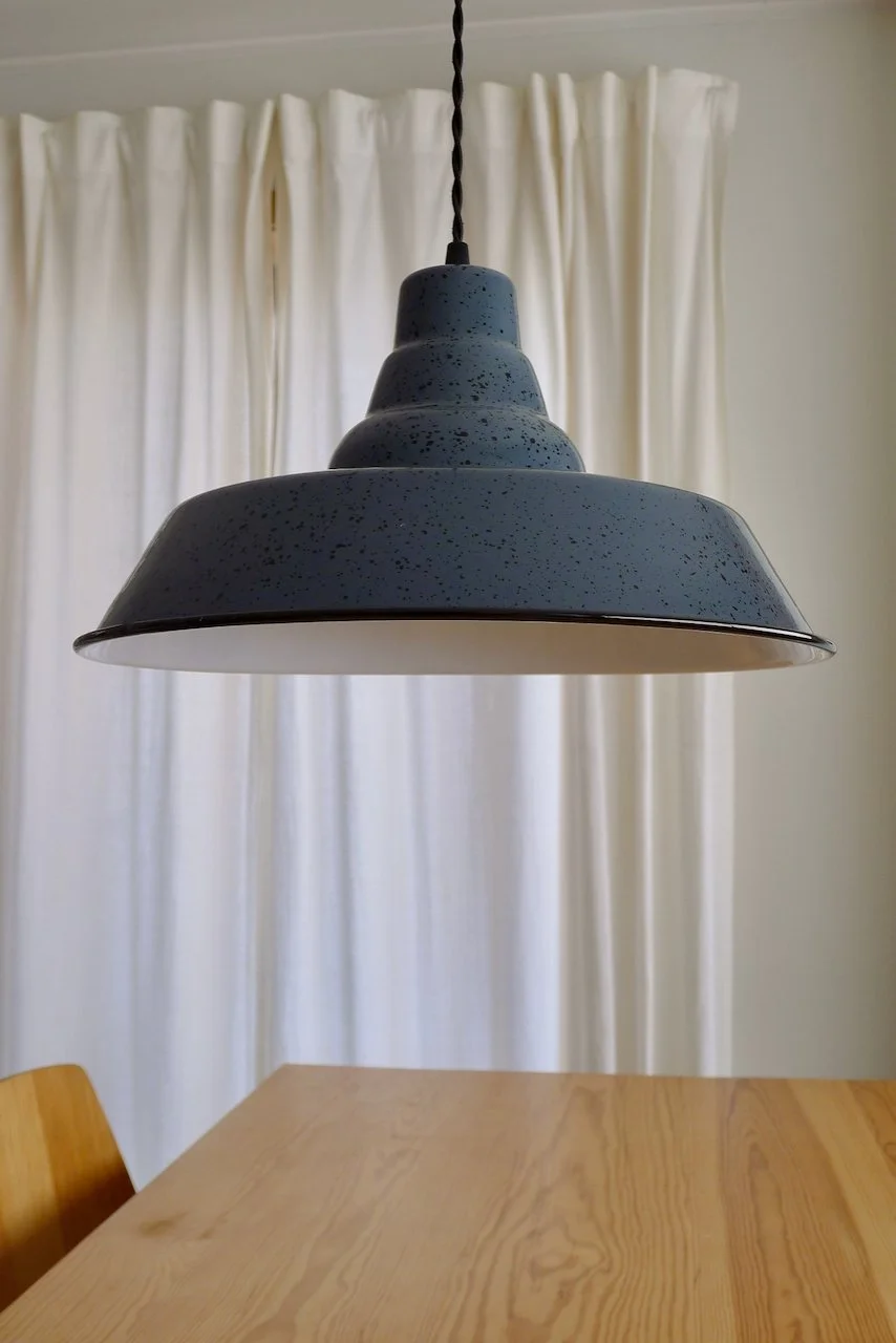 A dark-grey pendant light hanging above a wooden table with cream curtains in the background.
