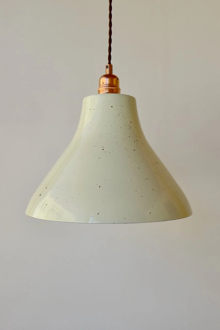 Close-up of a cream pendant lamp with a brass-colored metal cap, hanging from a dark-brown twisted cord against a plain light-colored wall.