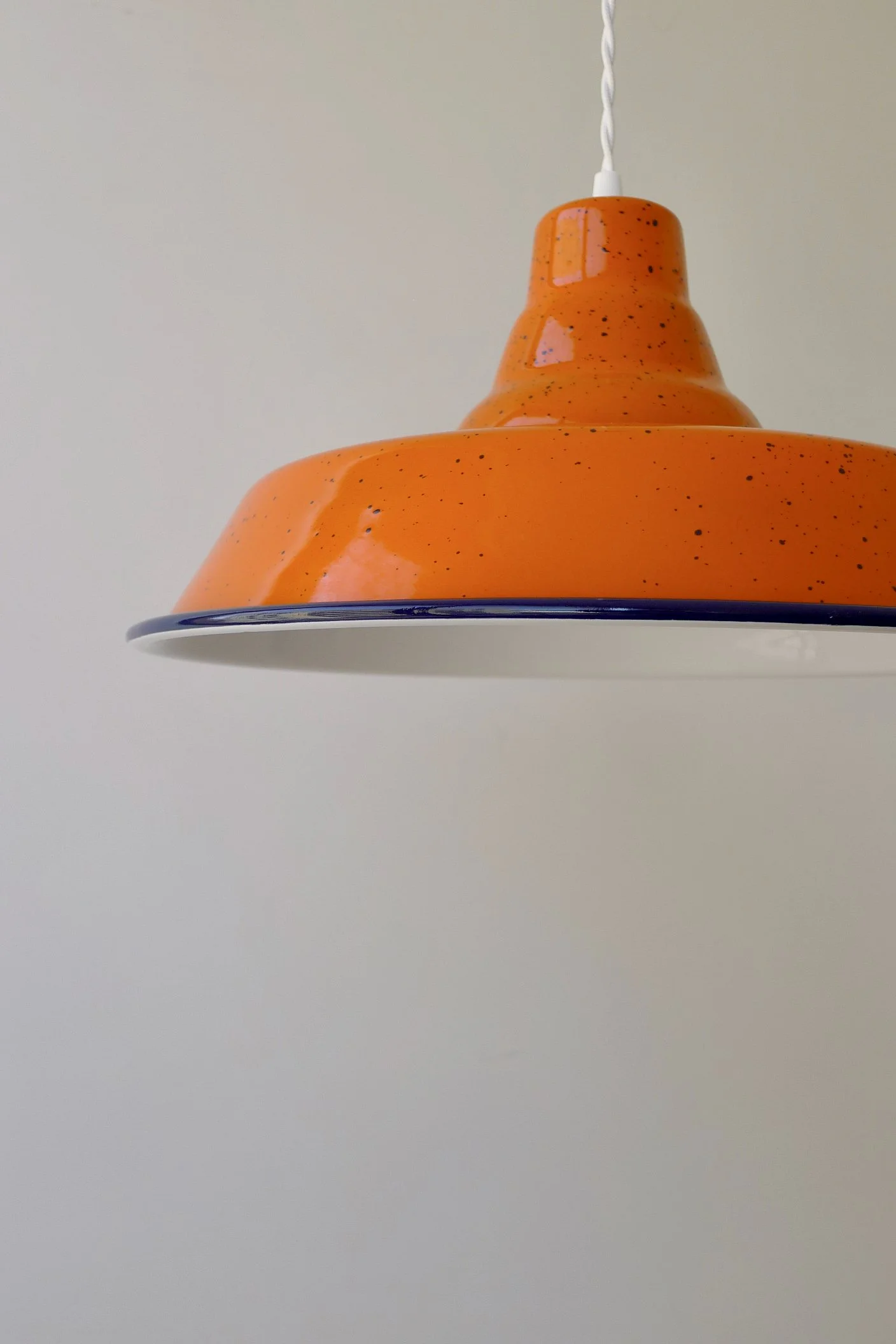 Close-up view of an orange enamel pendant lampshade with dark-blue speckles, hanging from a white twisted cord against a plain background.