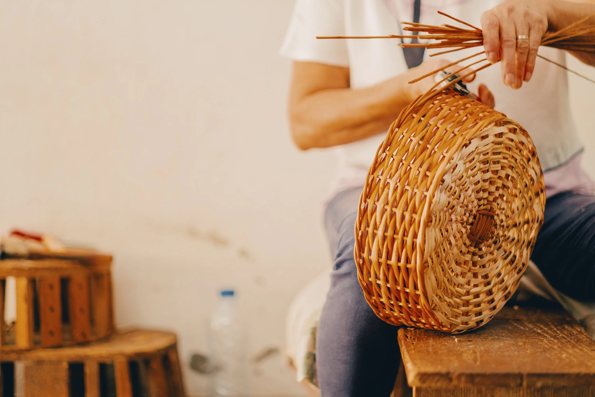 Person weaving a wicker basket, holding weaving sticks, in a workshop setting.