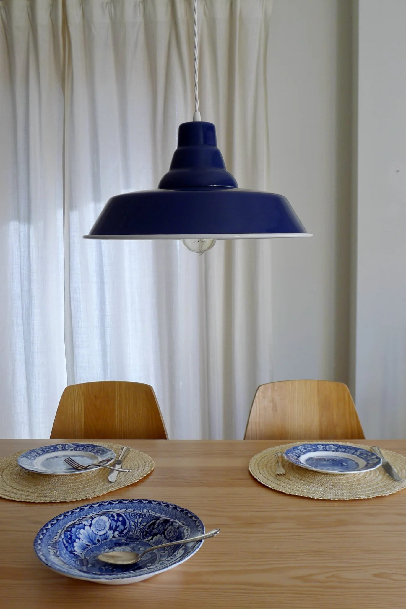 Dining table set with blue and white patterned plates, silverware, and woven placemats in a room with cream curtains and a navy-blue enamel pendant light.