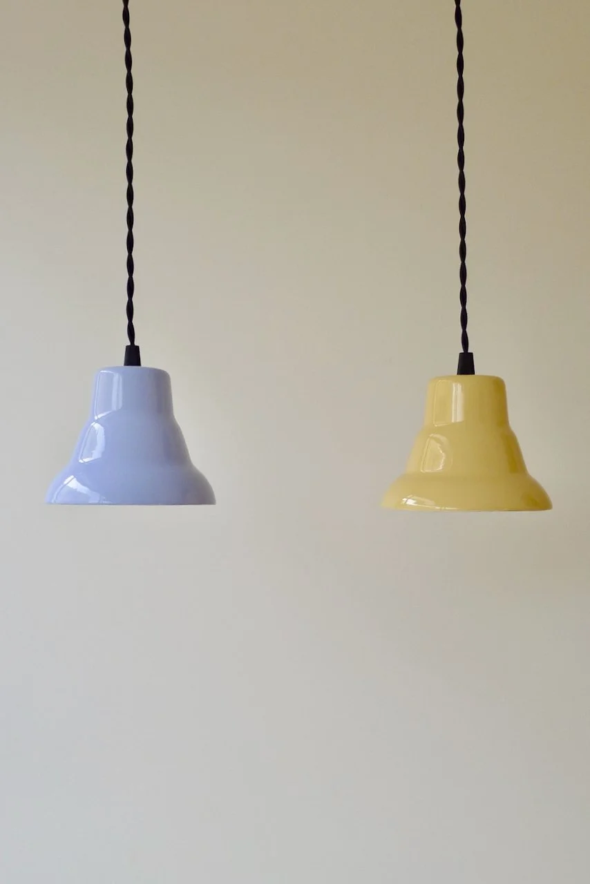 Two pendant lights, one lavender and one yellow, hanging from black twisted cords against a plain background.