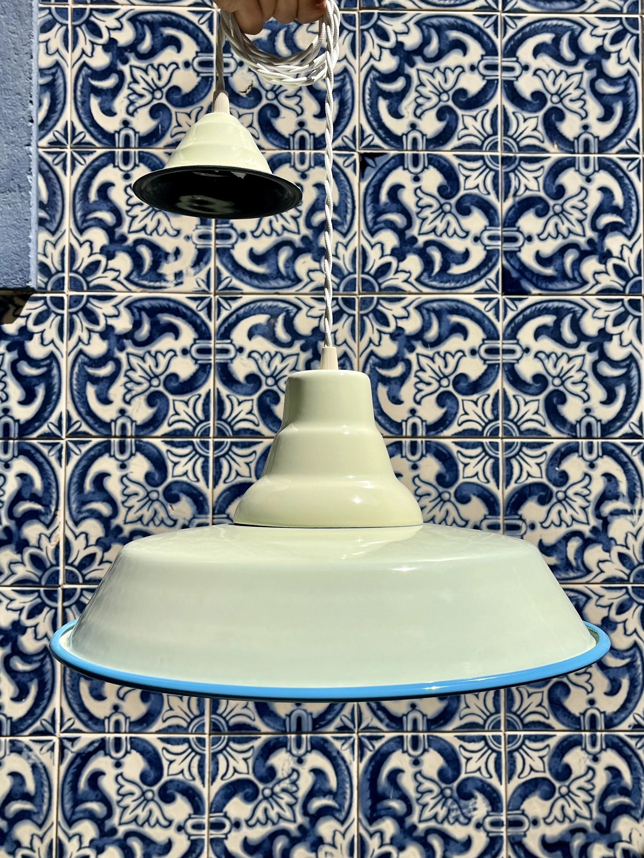 A white hanging pendant lamp with a black interior, hanging in front of a blue and white patterned tile wall.