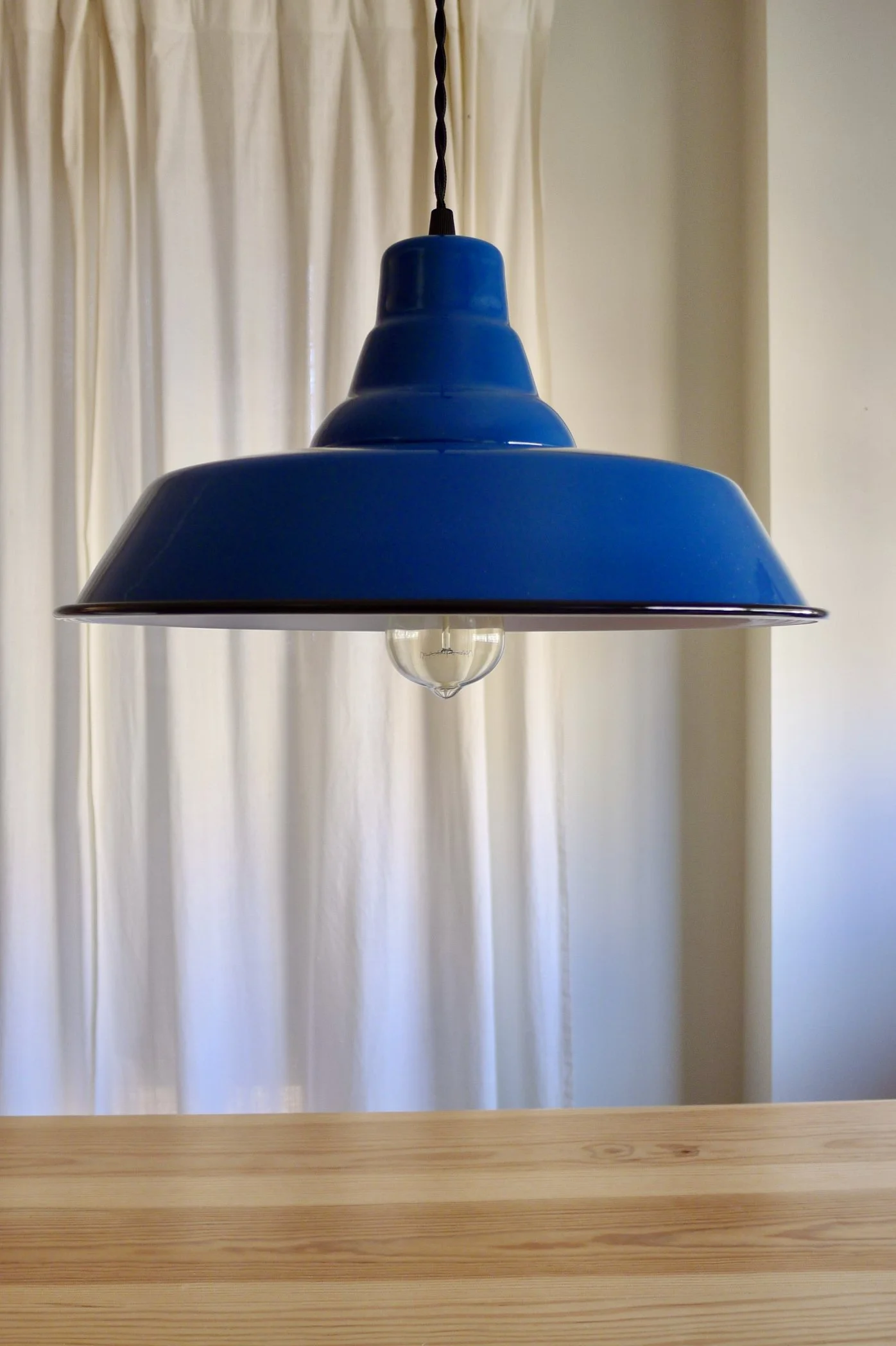 Blue pendant lampshade hanging above a wooden table with curtains in the background.