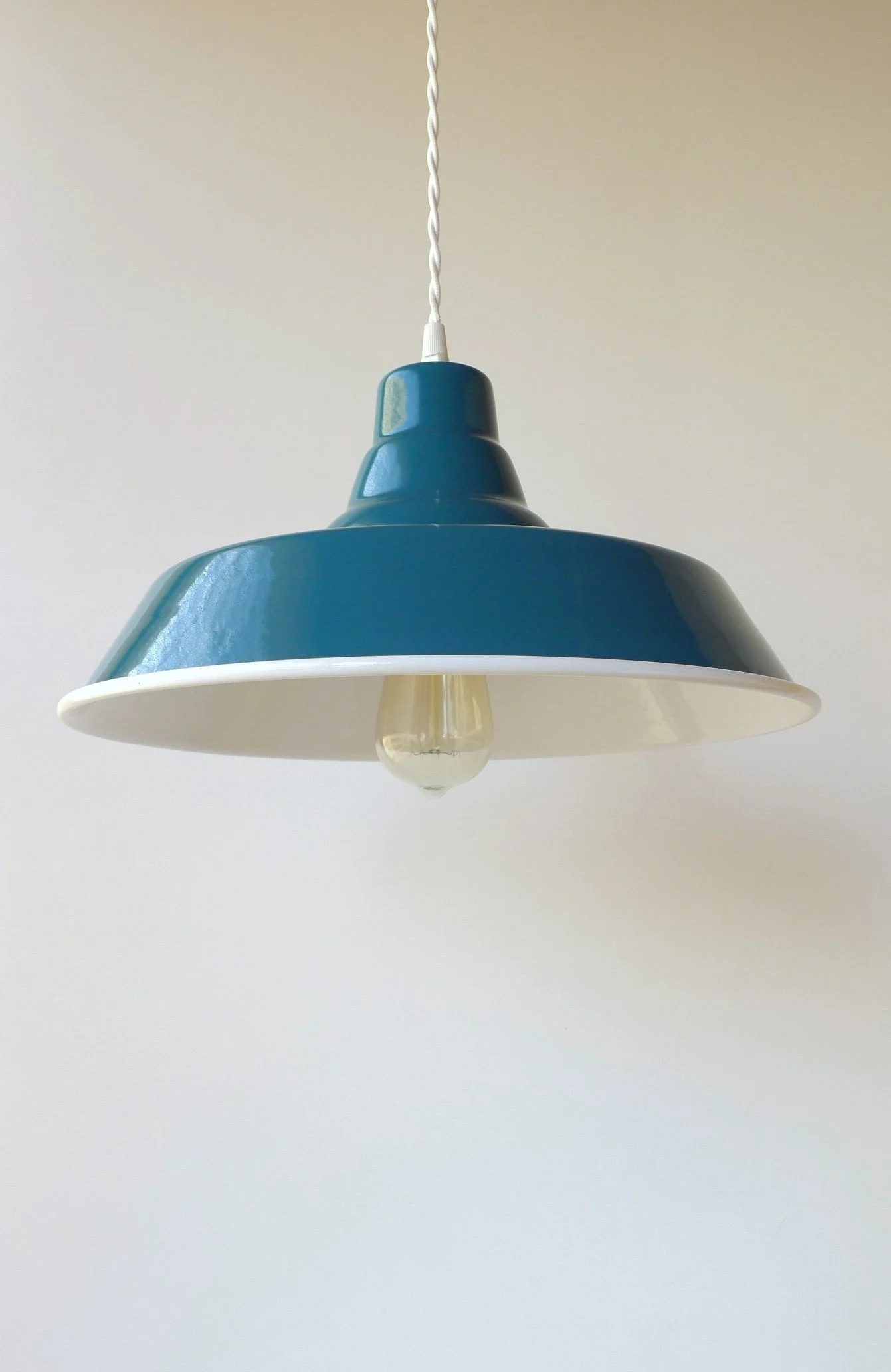 A blue pendant lampshade hanging from a white twisted cord with a white interior and a clear lightbulb.