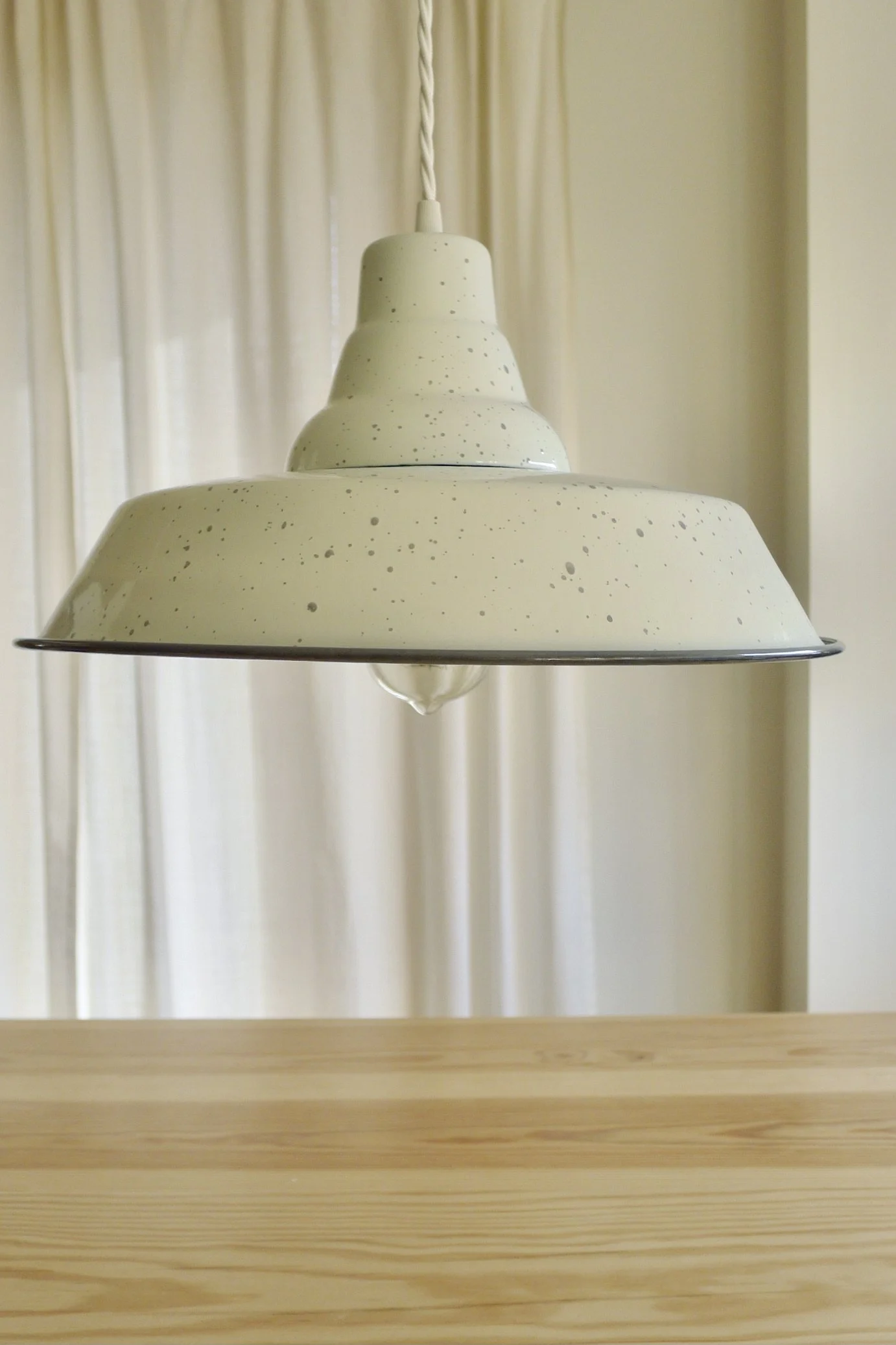 A cream-colored, speckled pendant light fixture hanging from a braided cord over a wooden surface, with curtains in the background.