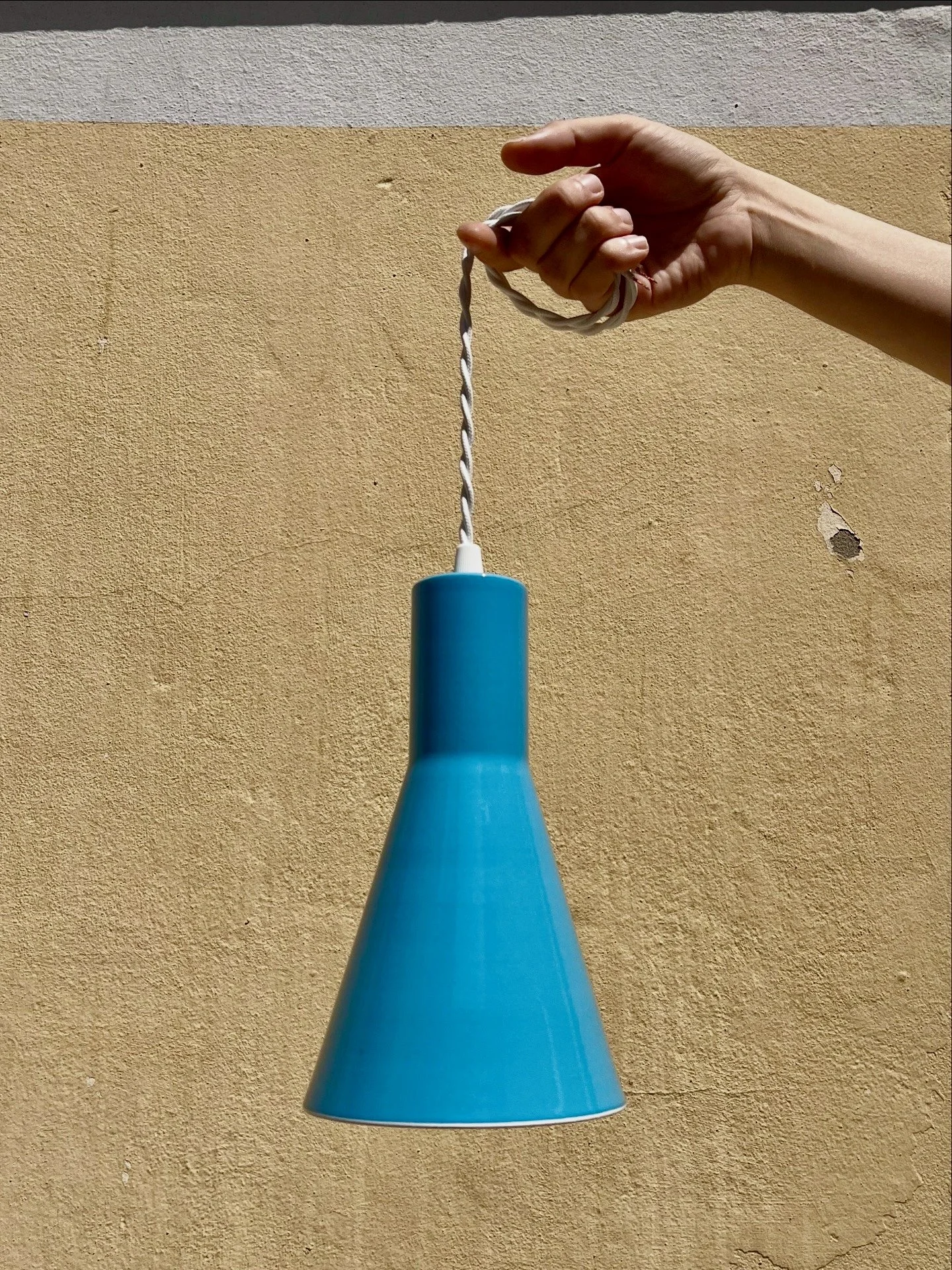 A blue cone-shaped hanging lamp with a twisted white fabric cord, against a two-tone textured wall painted in ochre and off-white.