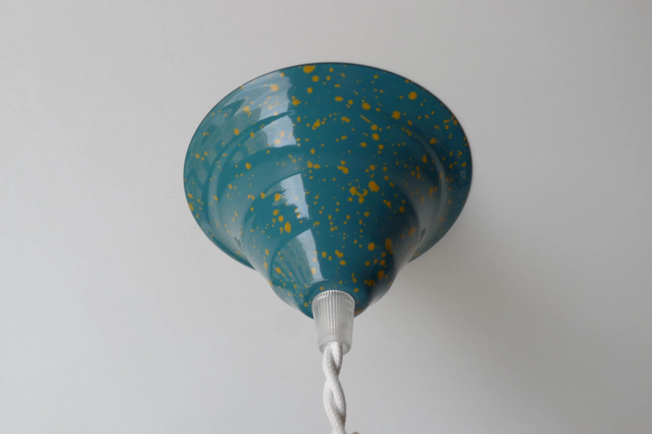 Green celing fixture with yellow splatter pattern, attached to a white string, against a plain white ceiling.