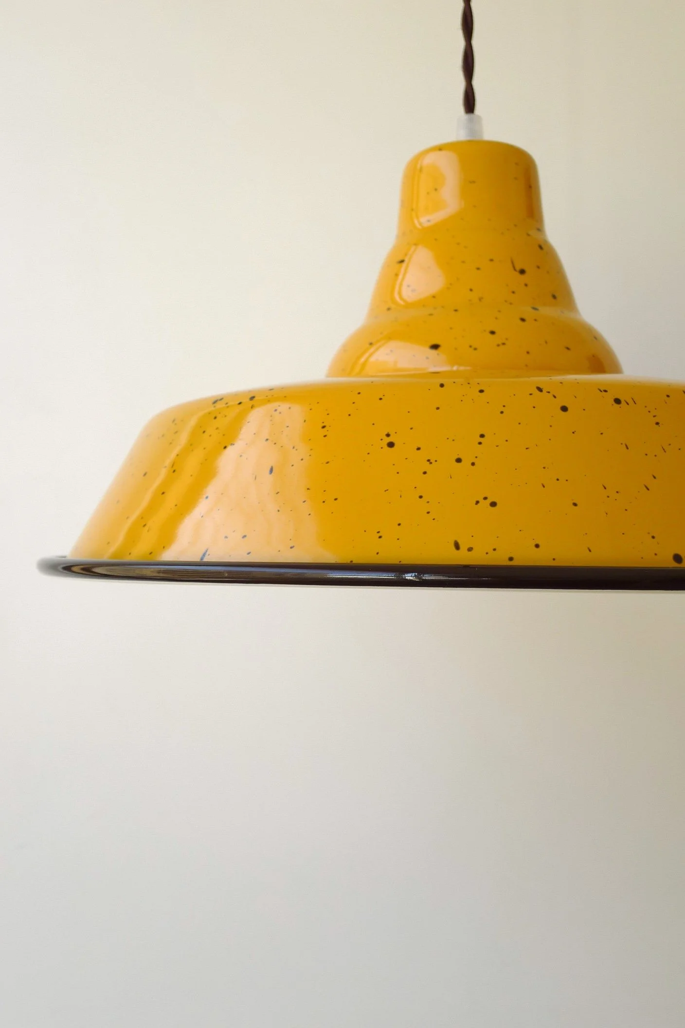 Close-up of a yellow, speckled enamel pendant light with a dark brown rim, hanging from a dark brown twisted cord against a plain white wall.