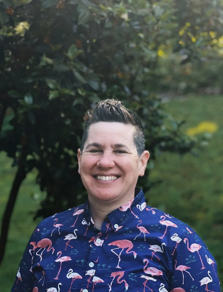 Person smiling wearing a shirt with a flamingo pattern, standing in front of green foliage.