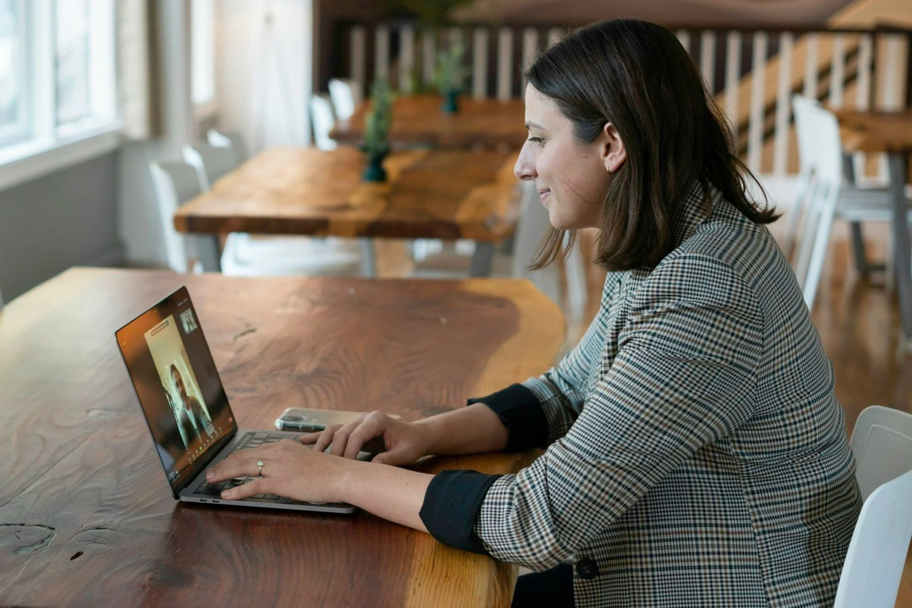 Woman in a plaid jacket sitting at a wooden table using a laptop for a video call.