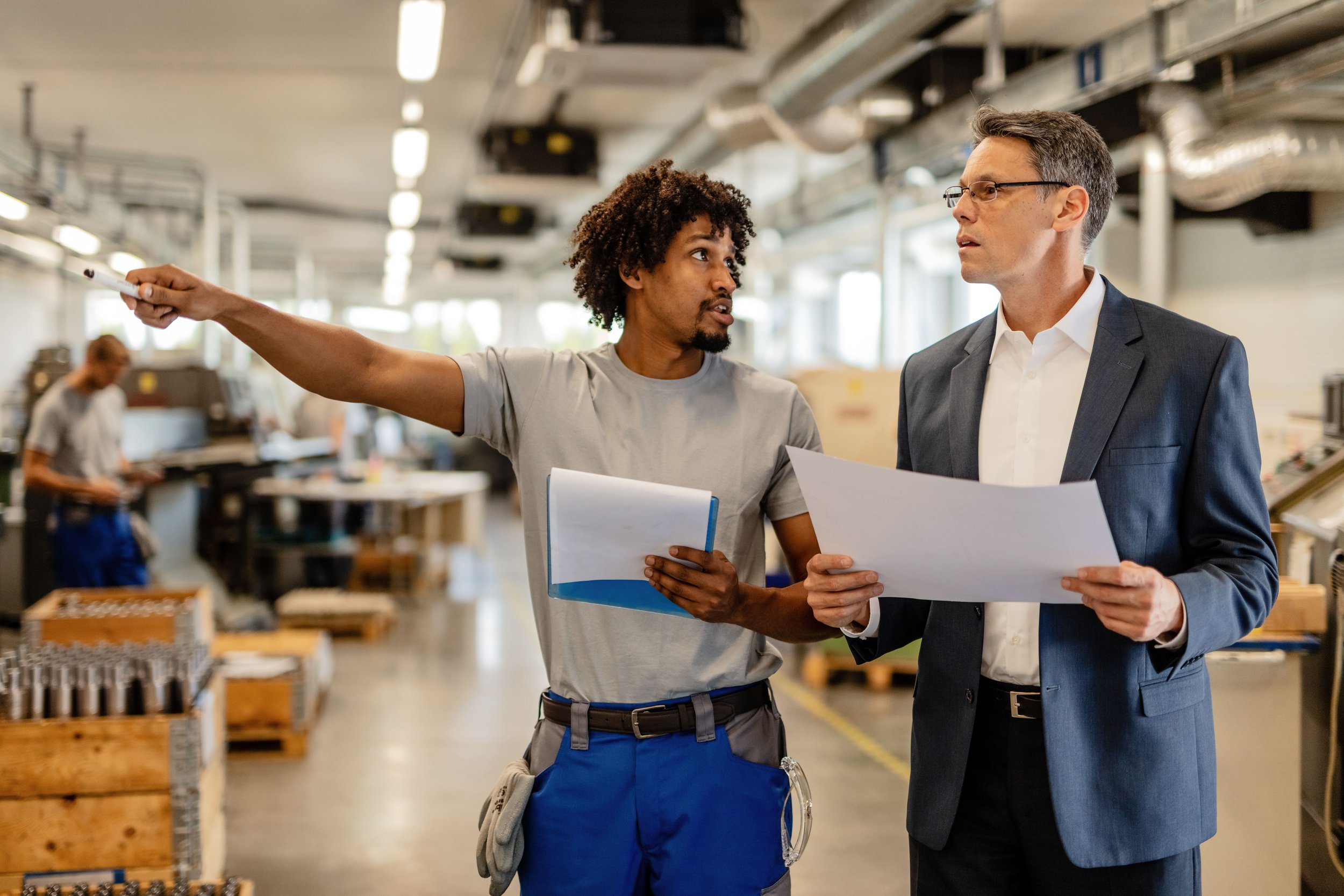 Two men, one in casual work clothes and the other in a suit, are having a discussion in an industrial or factory setting. The man in work clothes is pointing with a pencil, while the other holds papers and listens.