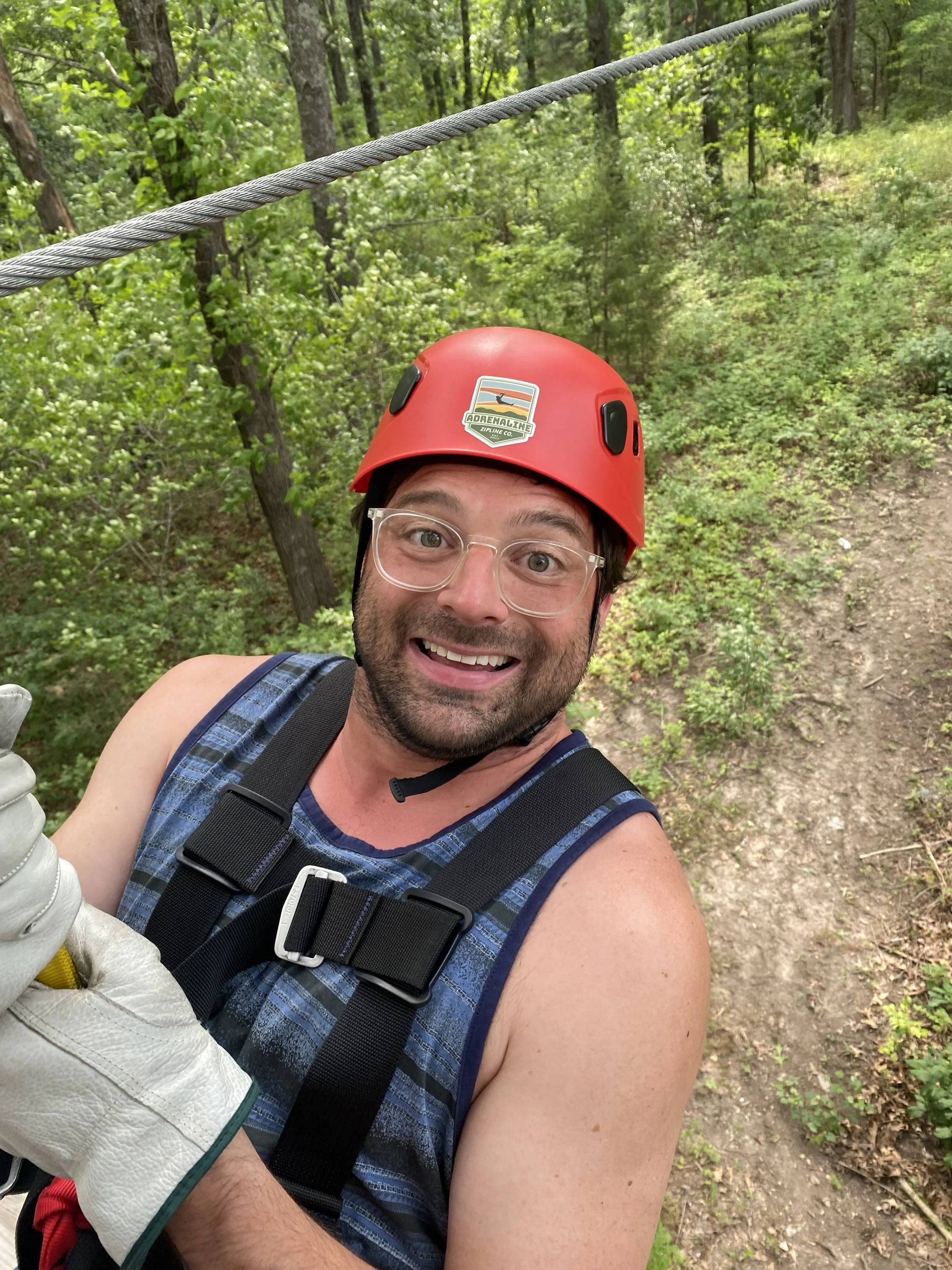 A man smiling and taking a selfie on a zip line, wearing a red helmet, glasses, a blue tank top, and a harness, surrounded by green trees in a forest.