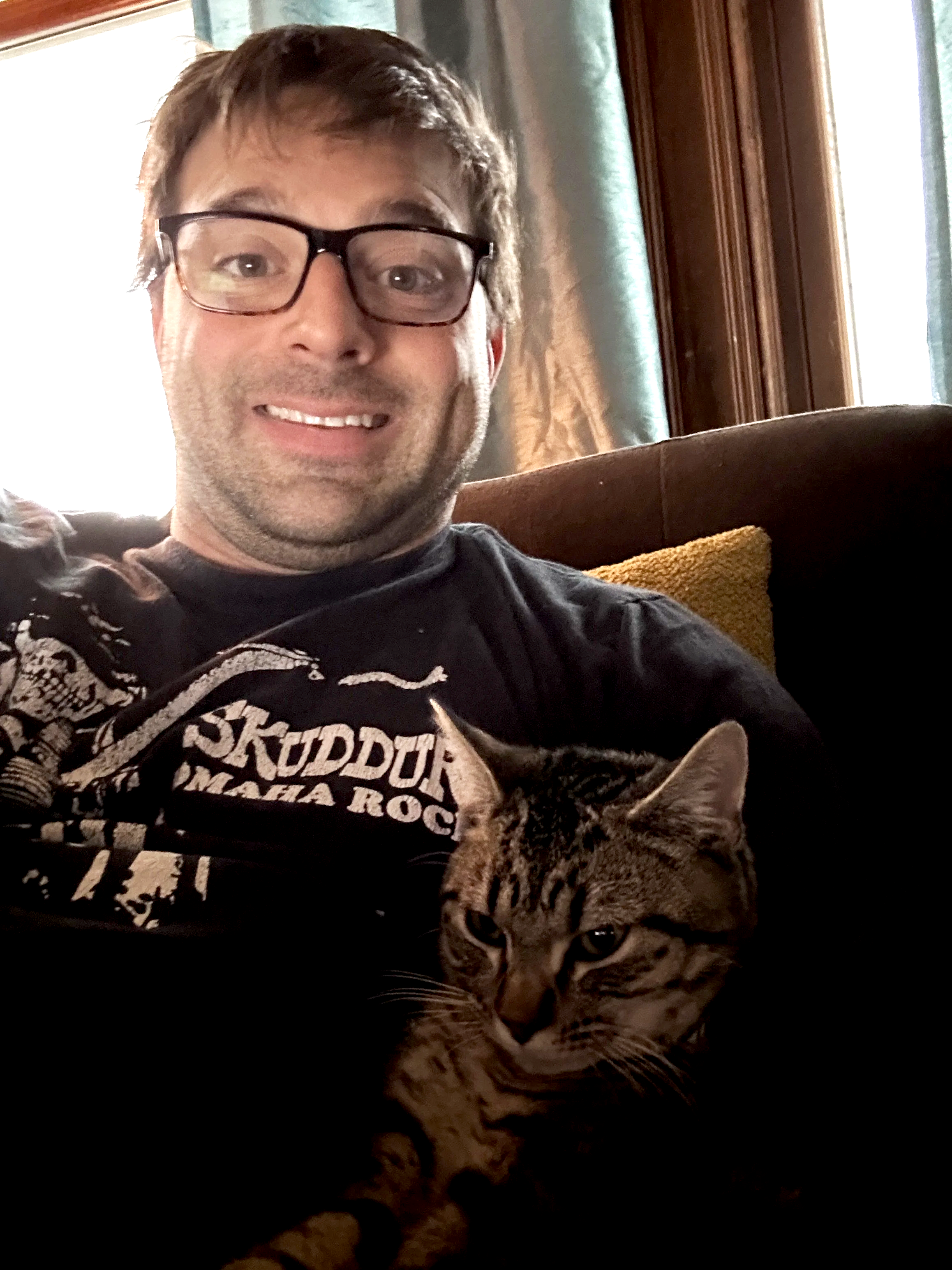A man wearing glasses smiling while sitting on a couch with a tabby cat.