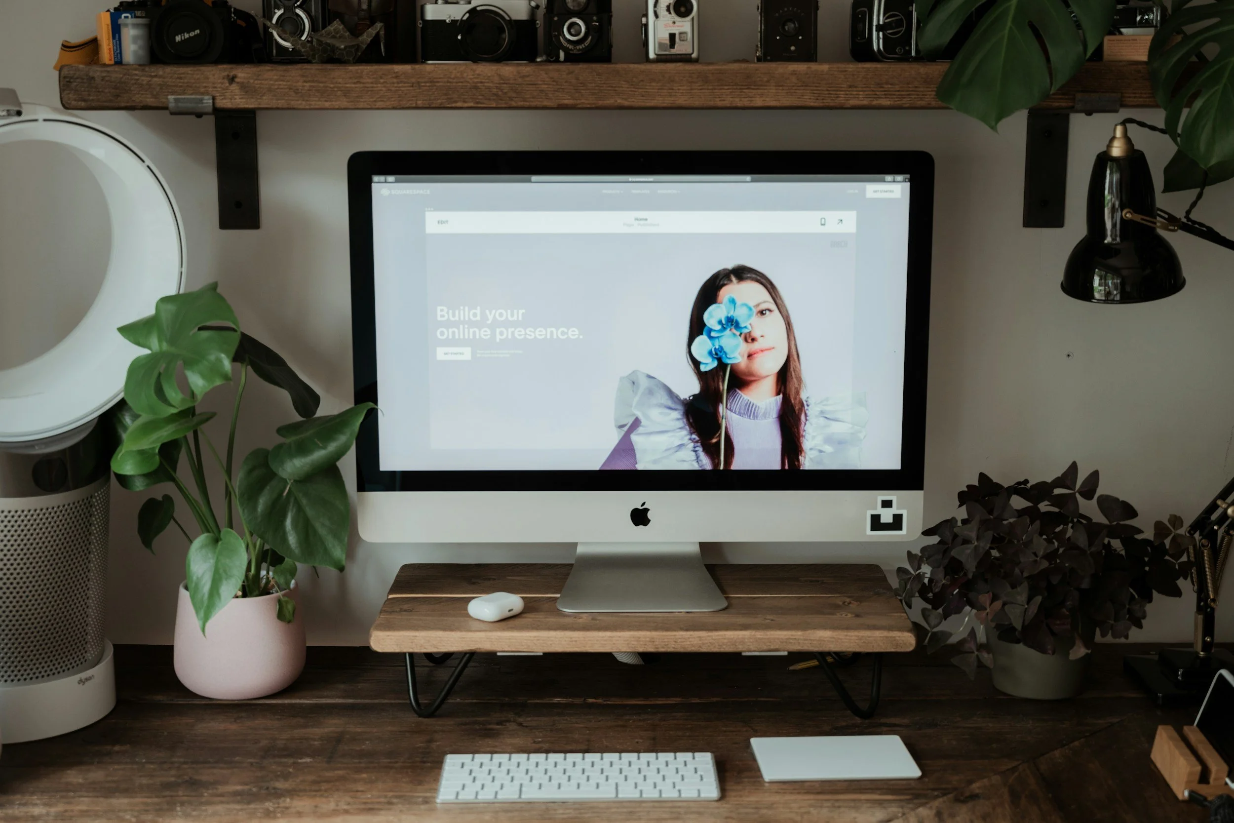 A computer workspace featuring an iMac showing a website with a woman holding an orchid in front of her face, surrounded by green plants, a keyboard, a mouse, and decorative plants on the desk, with a wooden shelf holding cameras above.