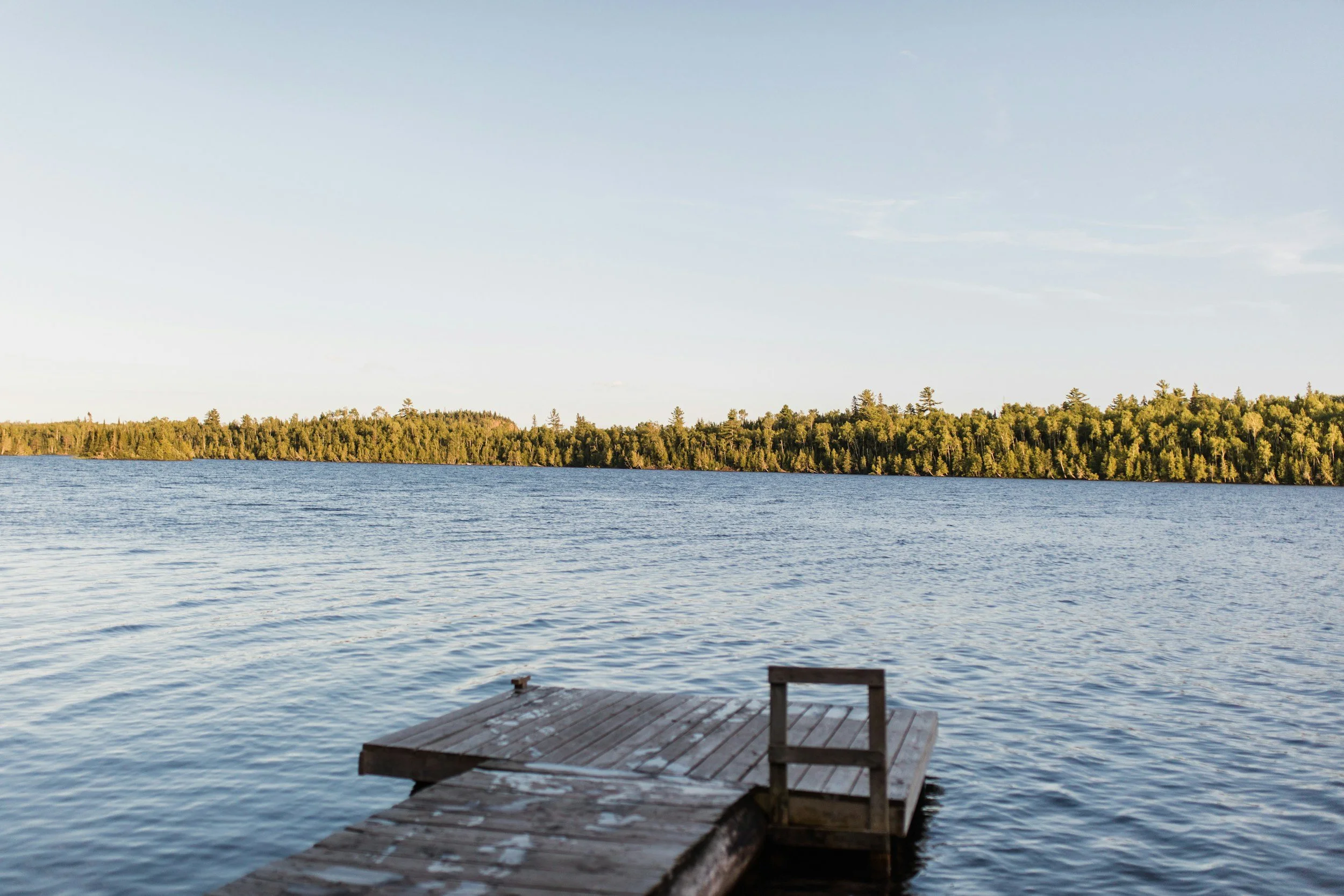 A wooden dock extends into a calm lake, with a distant treeline and clear sky in the background.