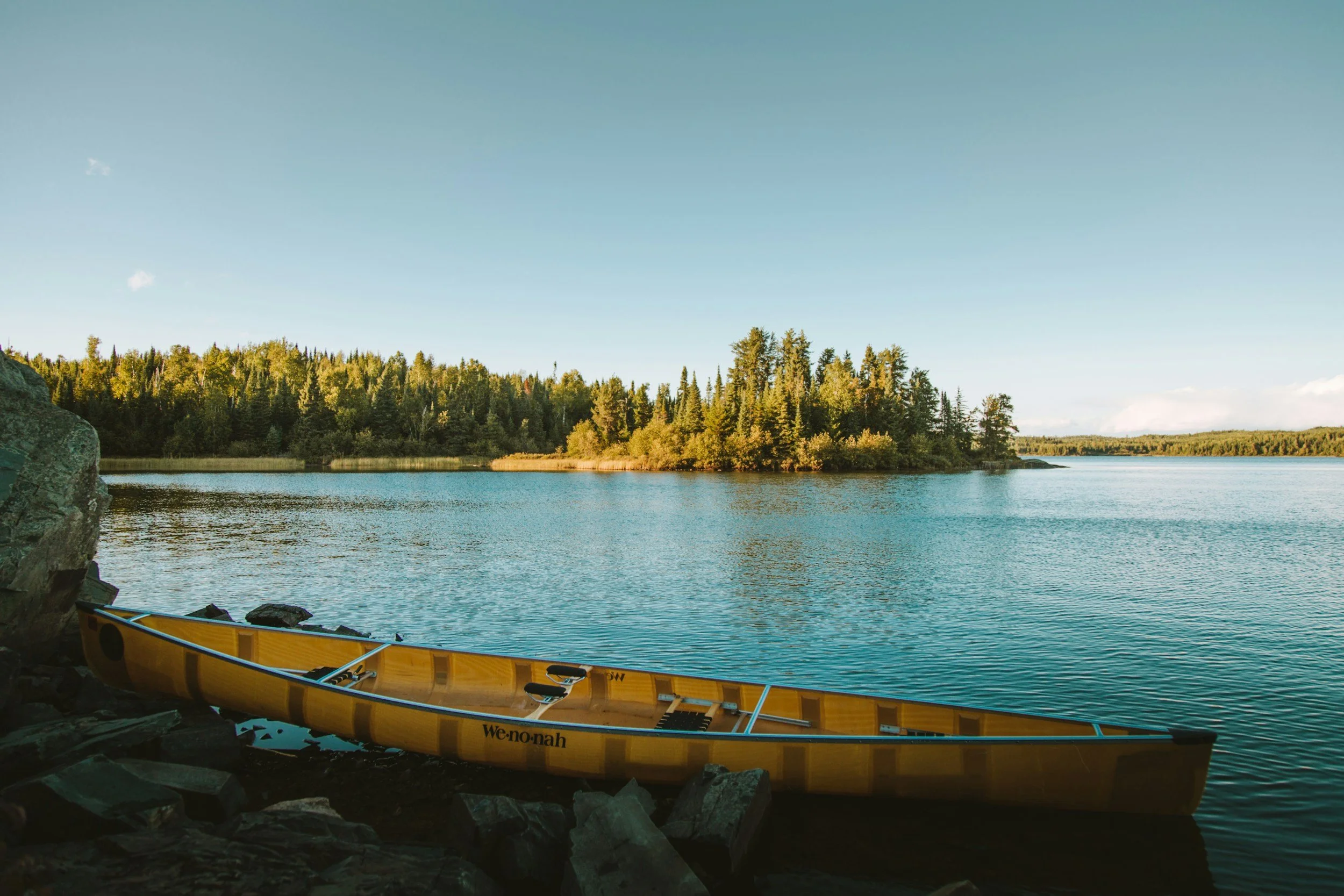 Yellow canoe resting on rocks by a calm lake with a forested island in the background under a clear sky.