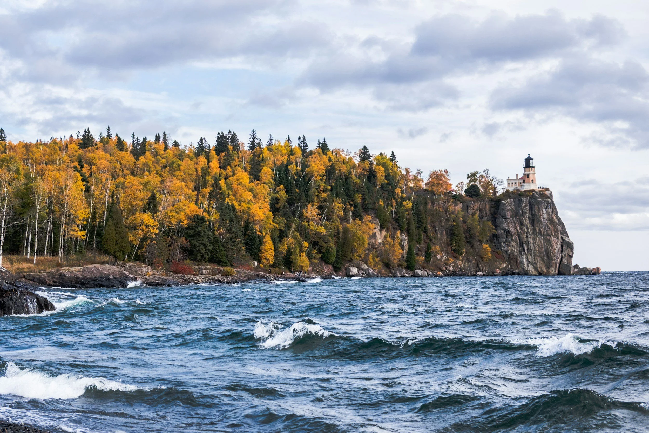 Lighthouse on a cliff overlooking a body of water with trees in fall colors and cloudy sky.