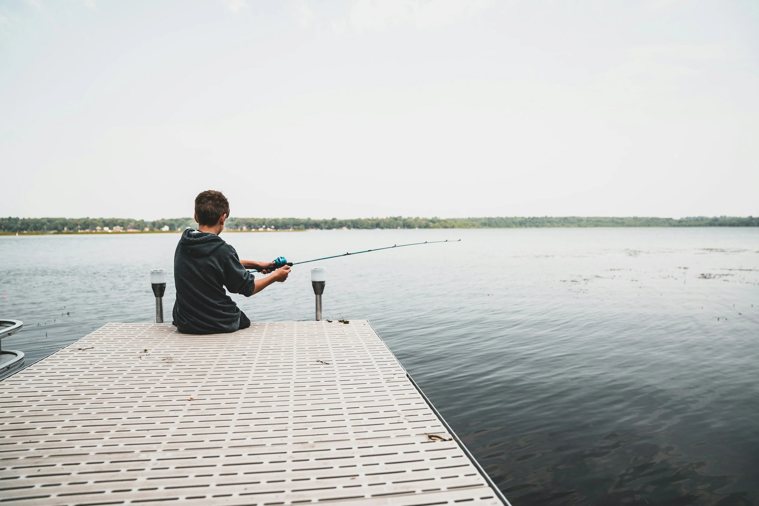 A boy sitting on a dock fishing in a lake during daytime.