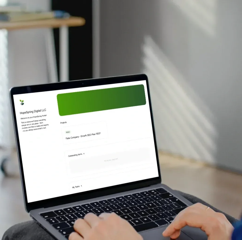 Laptop on a person's lap displaying a project management or organizational software called HopeSpring Digital LLC, showing a project with a green header, in a room with light coming through window blinds.