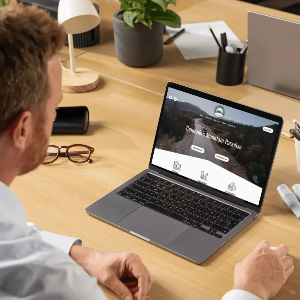 A person with glasses sitting at a wooden desk, viewing a laptop screen displaying a website about Colorado's Mountain Paradise, with a potted plant and office supplies on the desk.