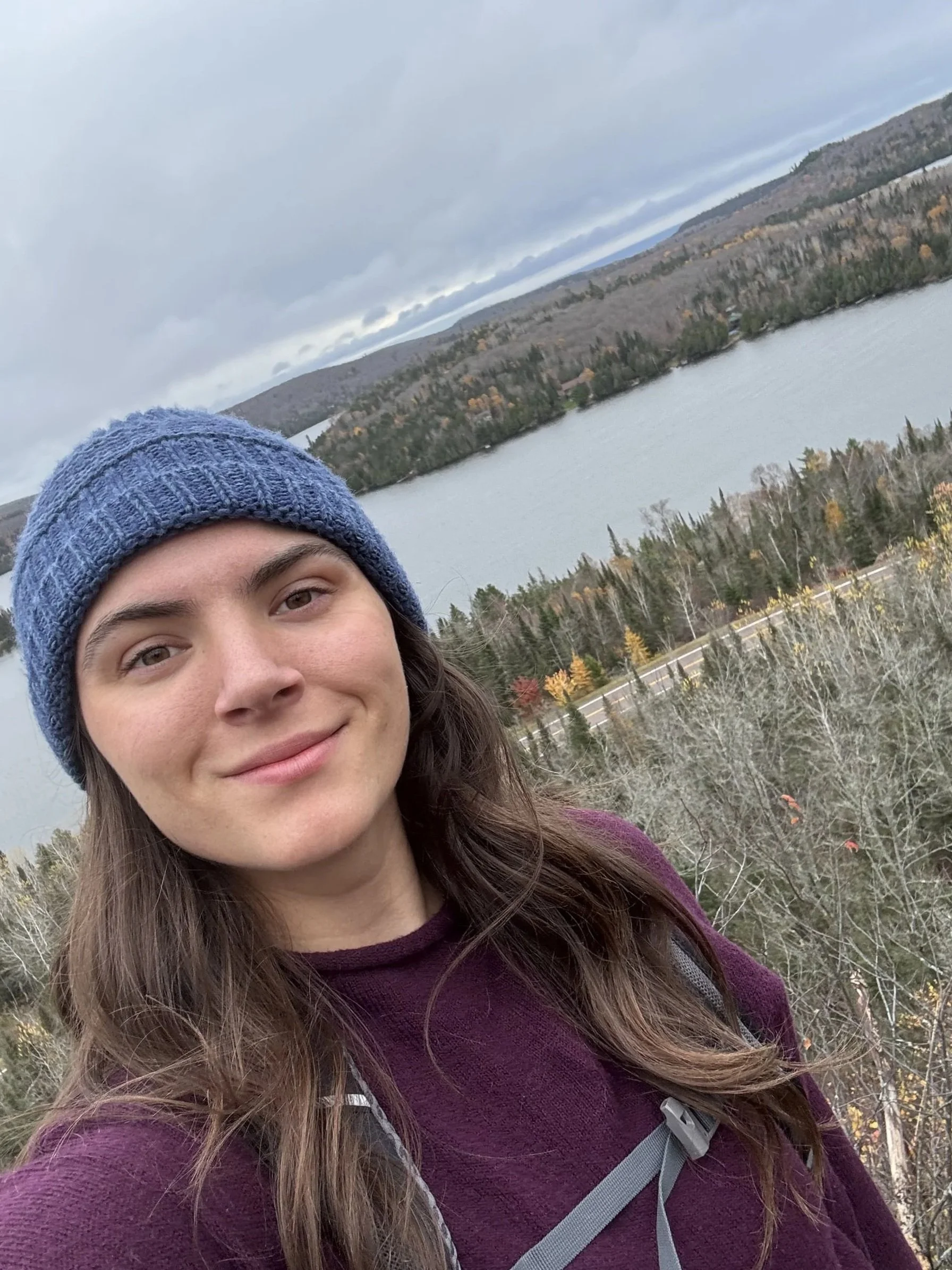 A young woman in a blue knit beanie and maroon jacket taking a selfie outdoors with a river, forested hills, and cloudy sky in the background.