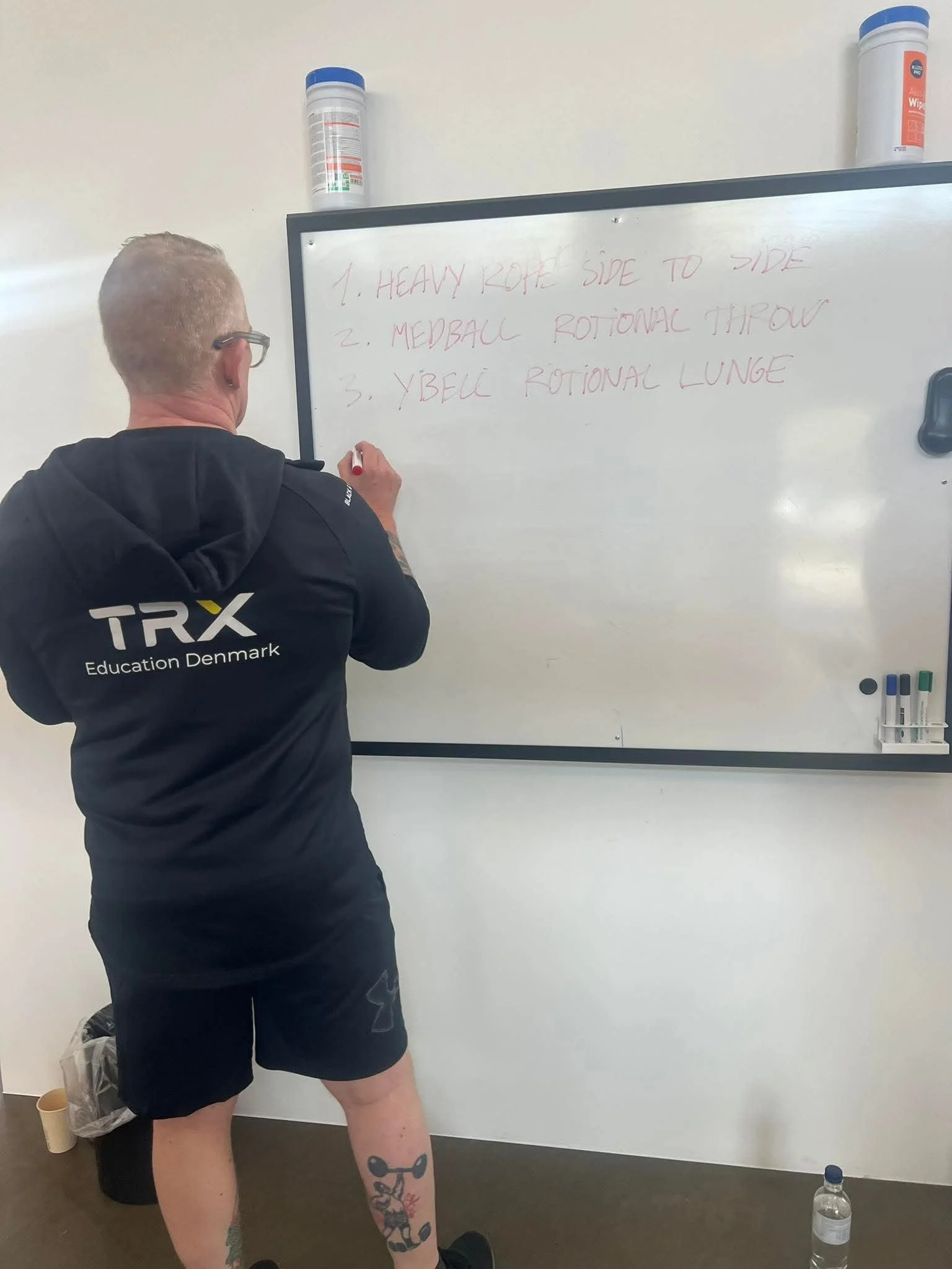 Person, der skriver på en hvid tavle med teksten 1. Heavy Rope Side To Side, 2. Medicine Rotary Thow, 3. Ybell Rotational Lunge.
