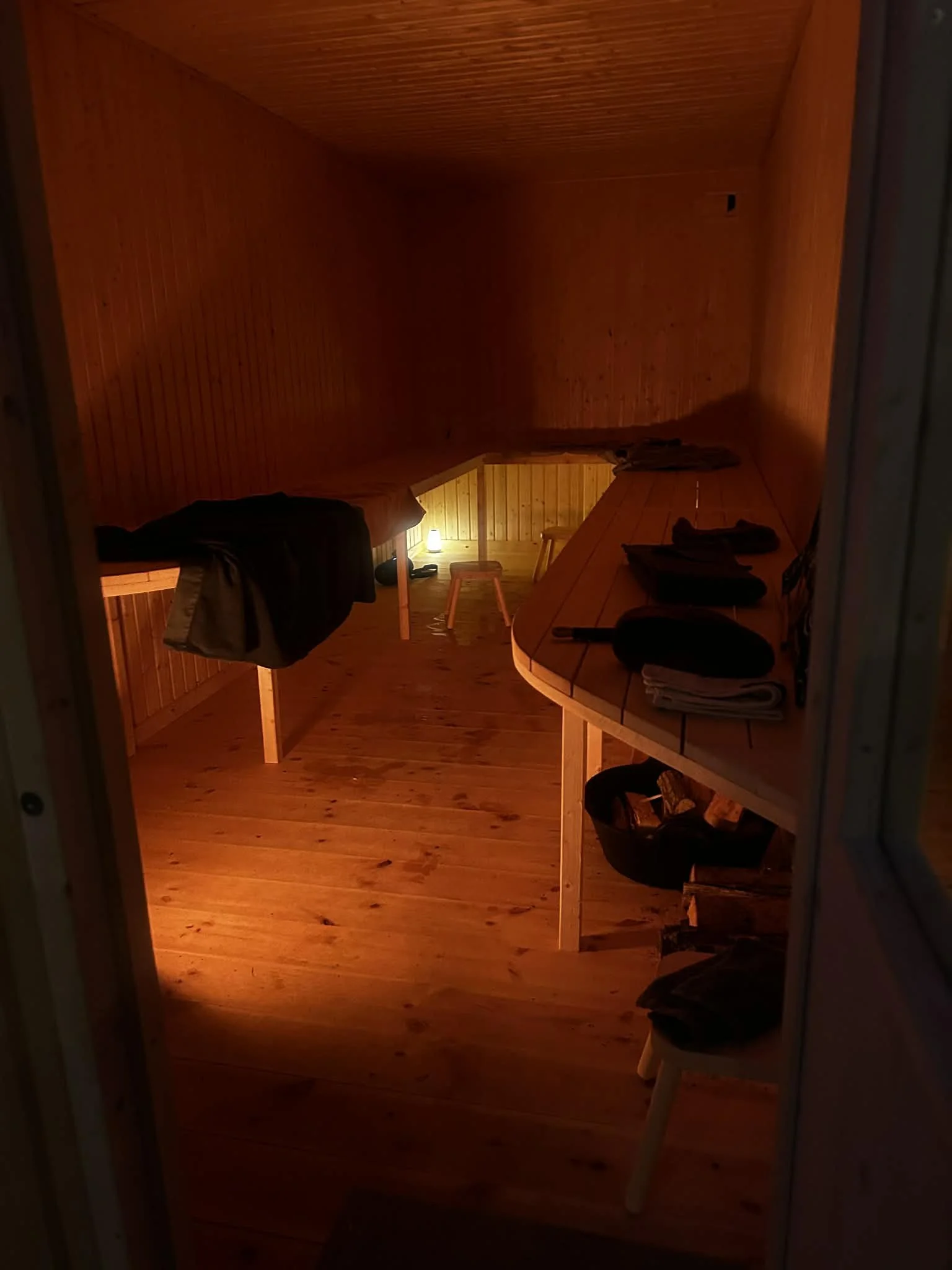 Indendørs rum med trævægge og trægulv, oplyst af et lille bordlampe, med tøj hængende på en stang og et bord med tøj og andre genstande.