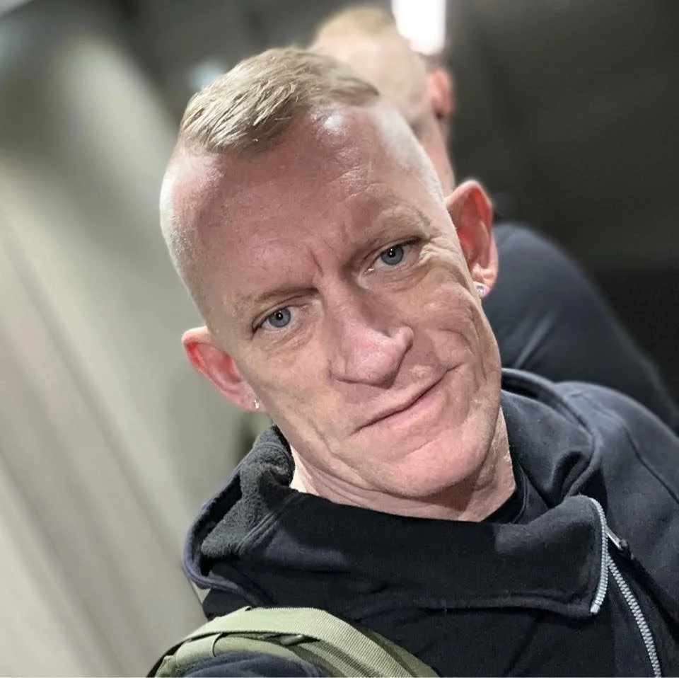 En mand med kort blond hår, blå øjne, let rynket ansigt og let bøjede ører, iført sort jakke, ser direkte i kameraet.