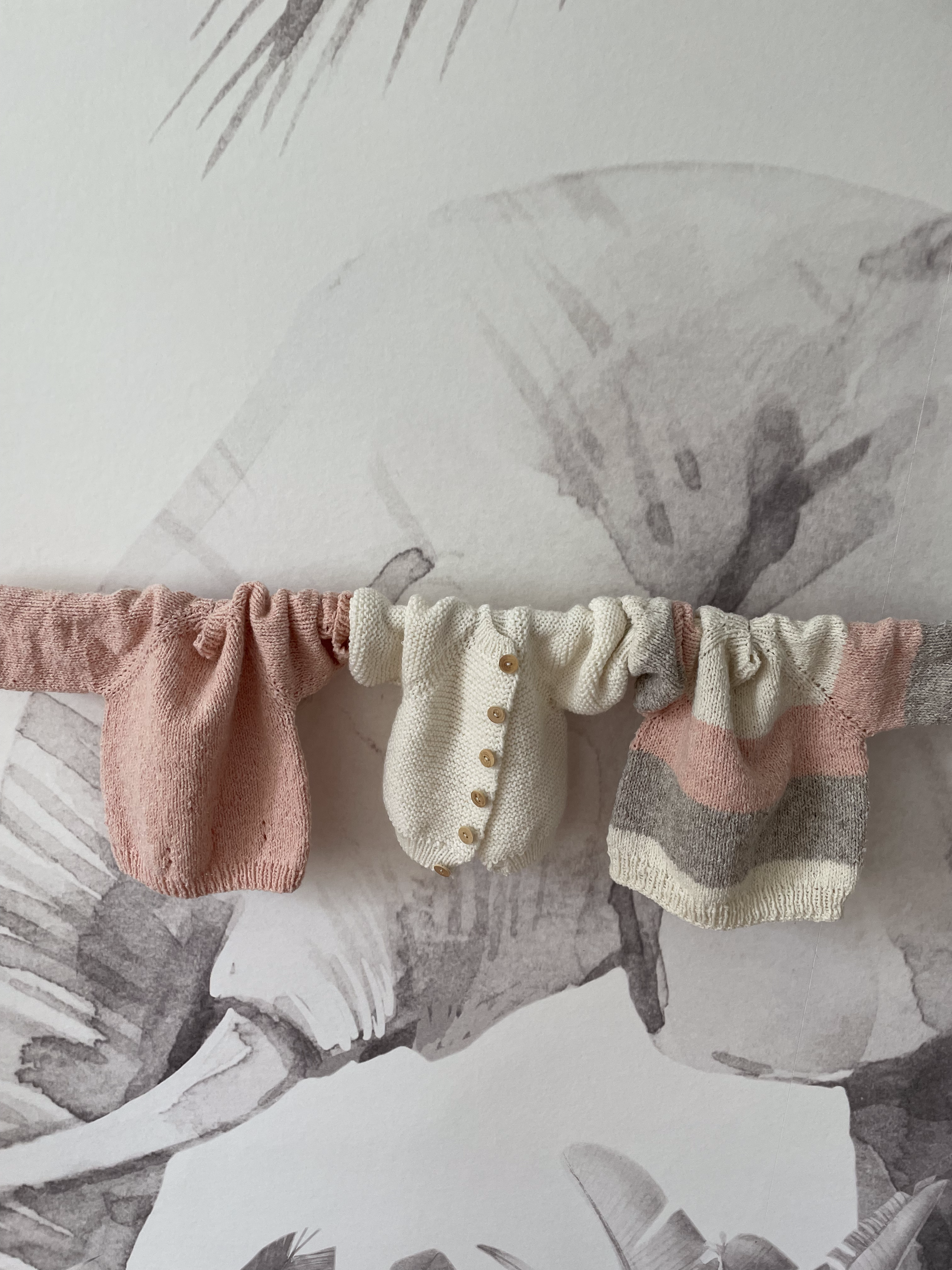 Deux petits chandails en coton, un rose et un à rayures gris, blanc et rose, avec un vêtement blanc entre eux, suspendus à une corde.
