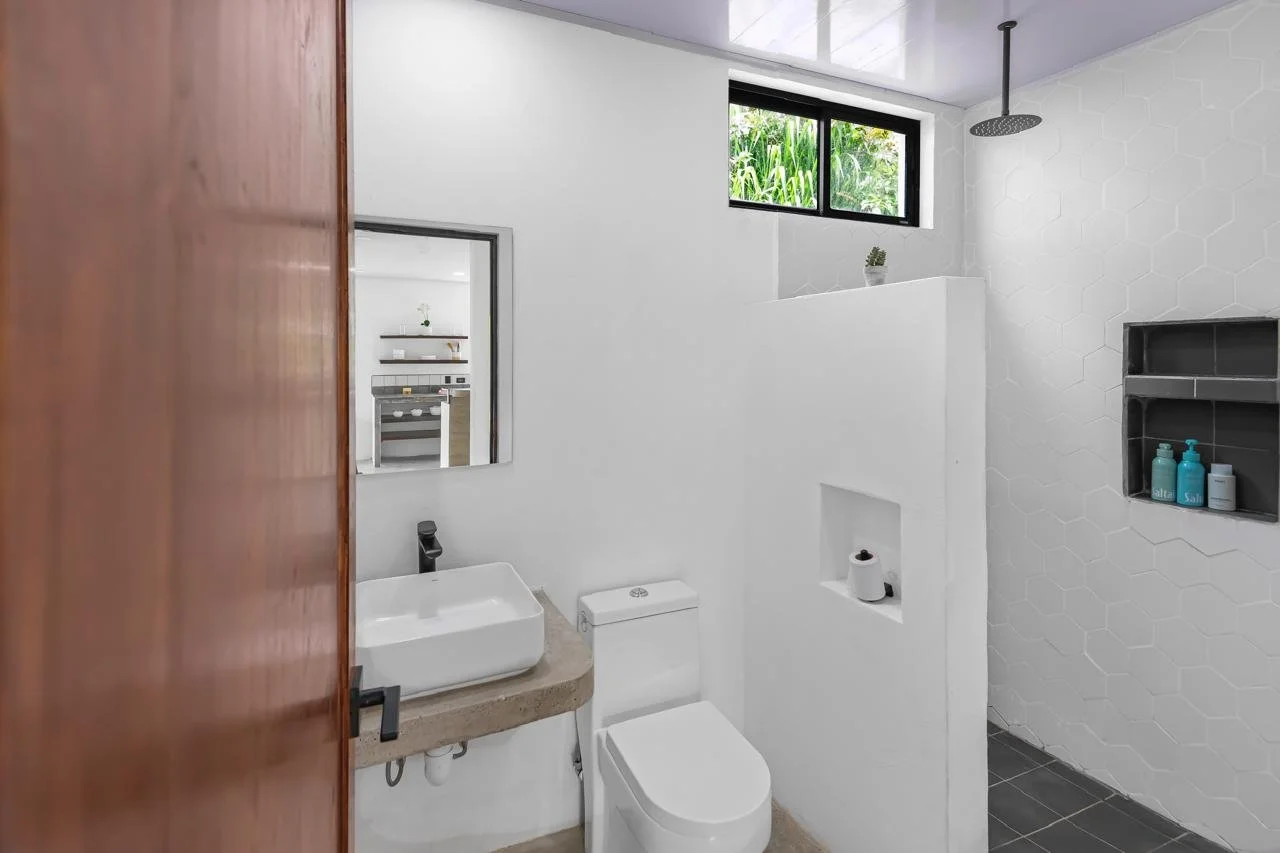 Modern bathroom with white walls, a small rectangular sink on a concrete countertop, a white toilet, a mirror, and a window with greenery outside. Shower area with a rain shower head and built-in shelves with various toiletries.