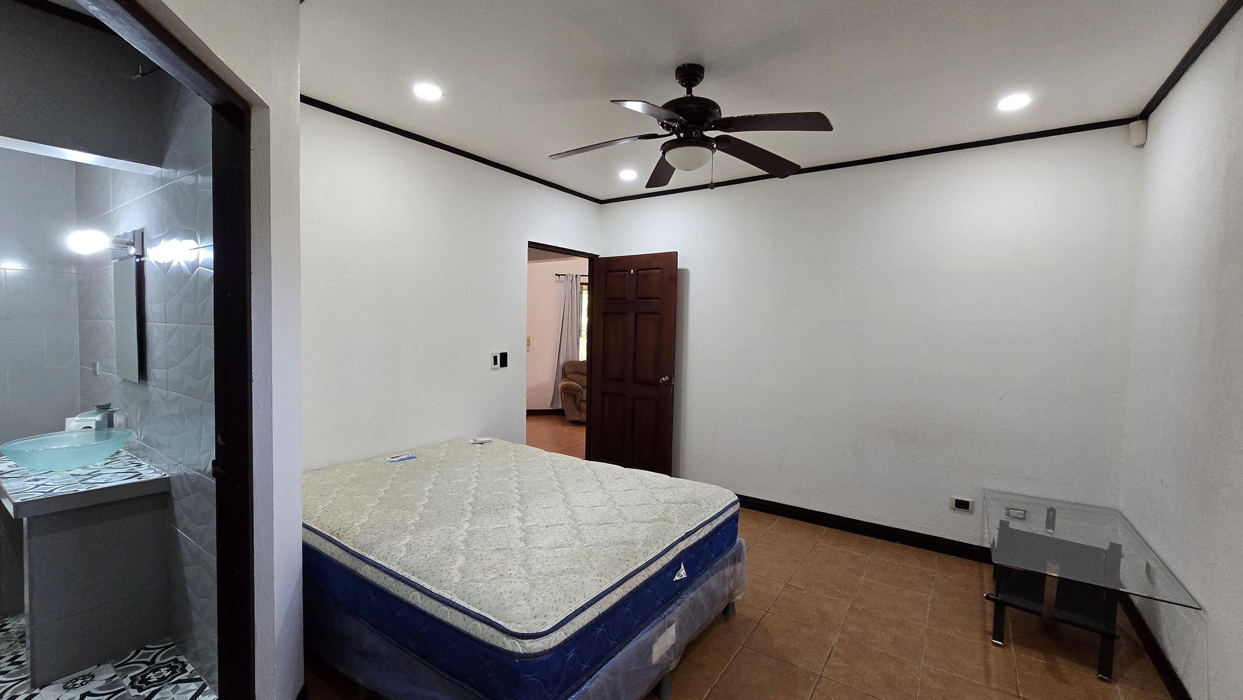 Empty bedroom with a bed, a ceiling fan, a glass table, and a door leading to a living room with a recliner chair.