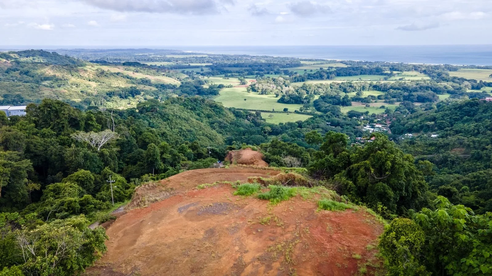 View from a hilltop overlooking lush green forest, rolling farmland, and distant ocean on a partly cloudy day.