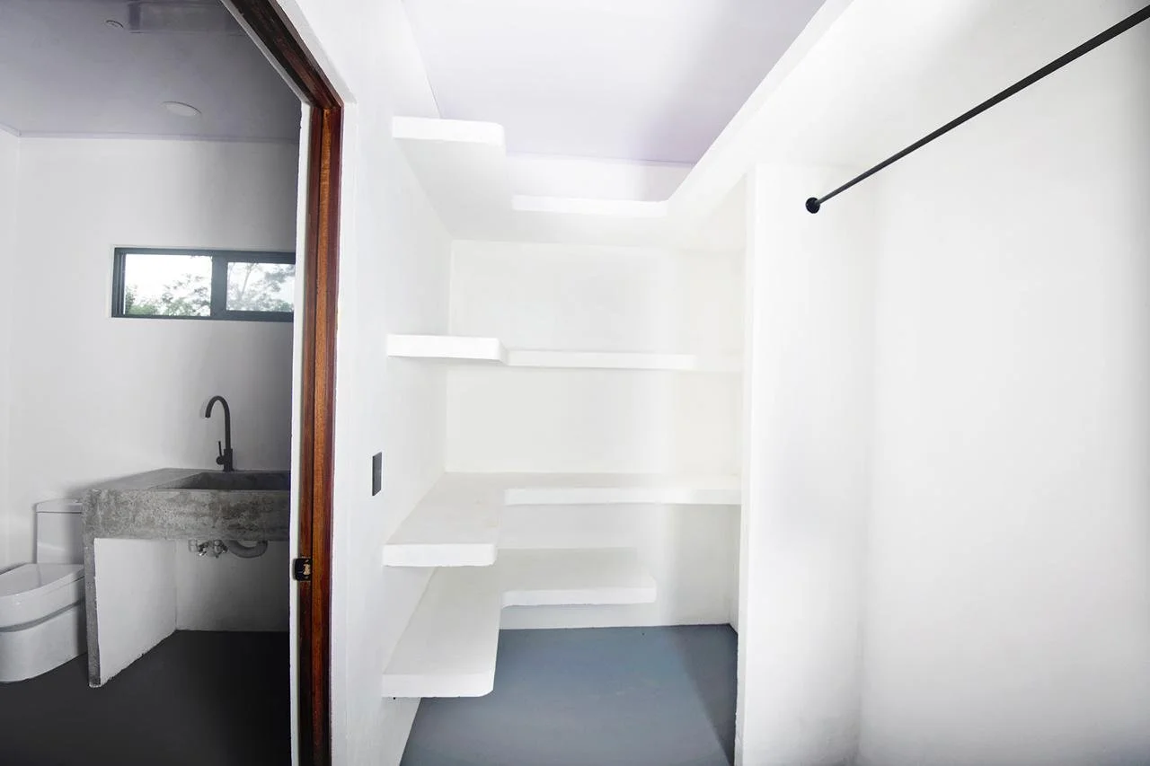 Empty white room with built-in white shelves on the wall and a dark-colored rod for hanging clothes. There is a small bathroom visible through an open sliding door, with a window, a sink, and a toilet.