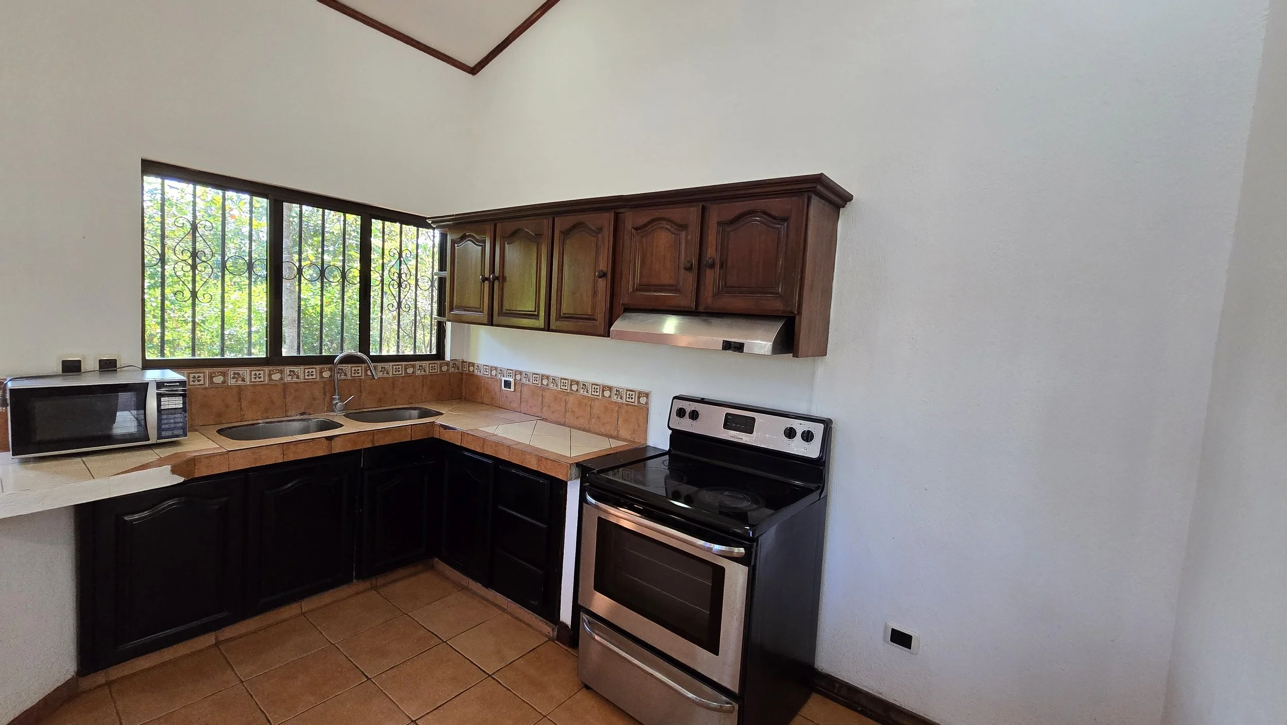 Kitchen with wooden upper cabinets, black lower cabinets, a stainless steel stove, a microwave, a tile countertop, a window with decorative iron bars, and terracotta floor tiles.